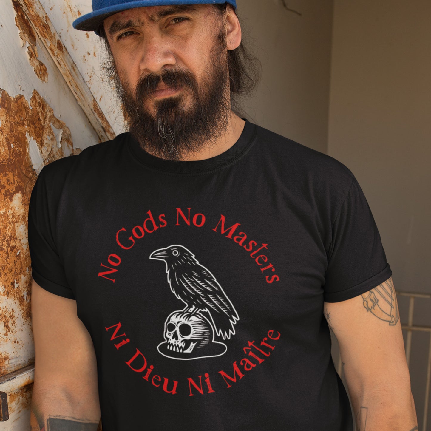 Model wearing black unisex t-shirt with raven and skull illustration, circular red text reading “NO GODS NO MASTERS” and “NI DIEU NI MAÎTRE,” casual indoor setting.