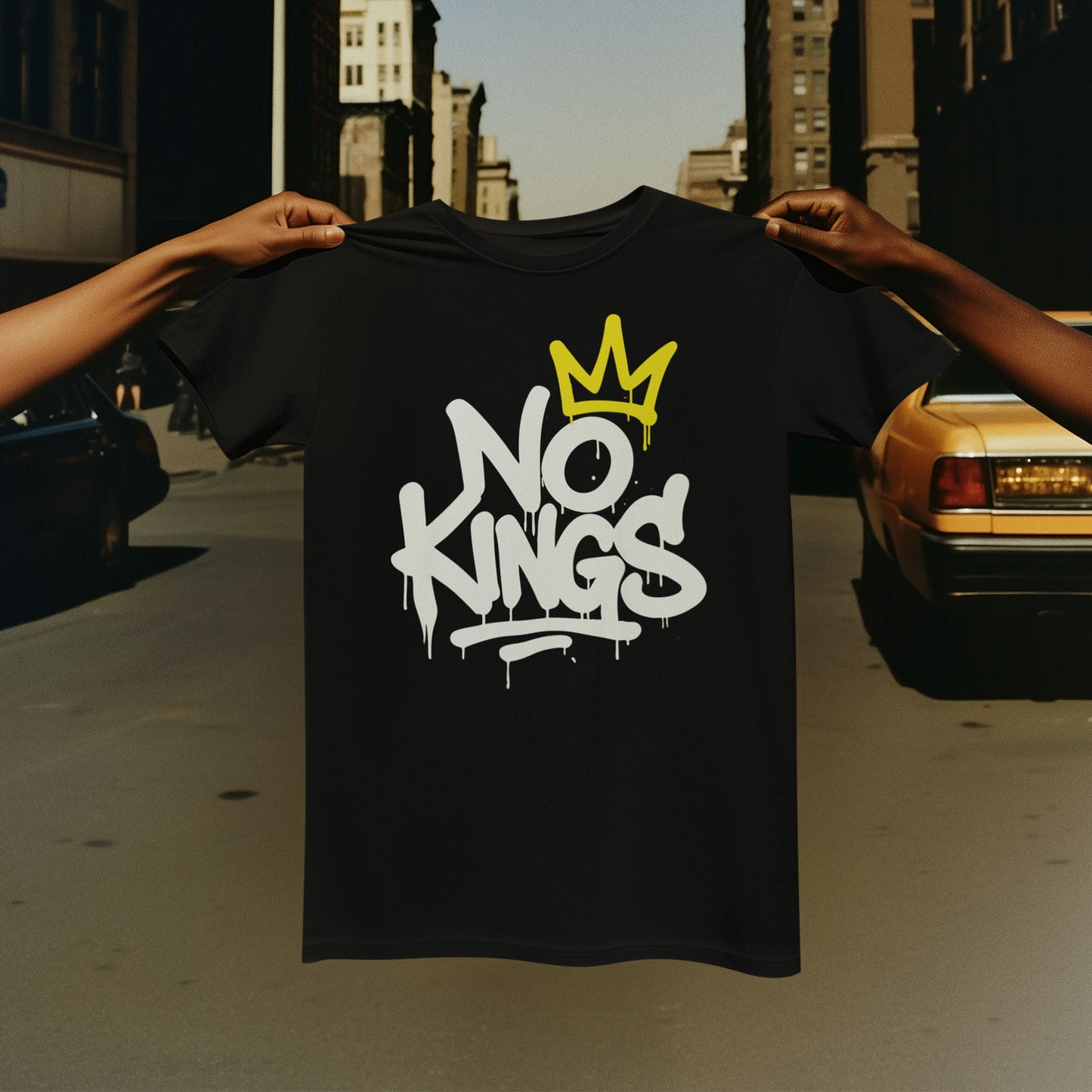 Person holding up a black “No Kings” t-shirt with white graffiti lettering and yellow crown detail against a city background. Anti-MAGA Merch designed by Blue Wave USA.
