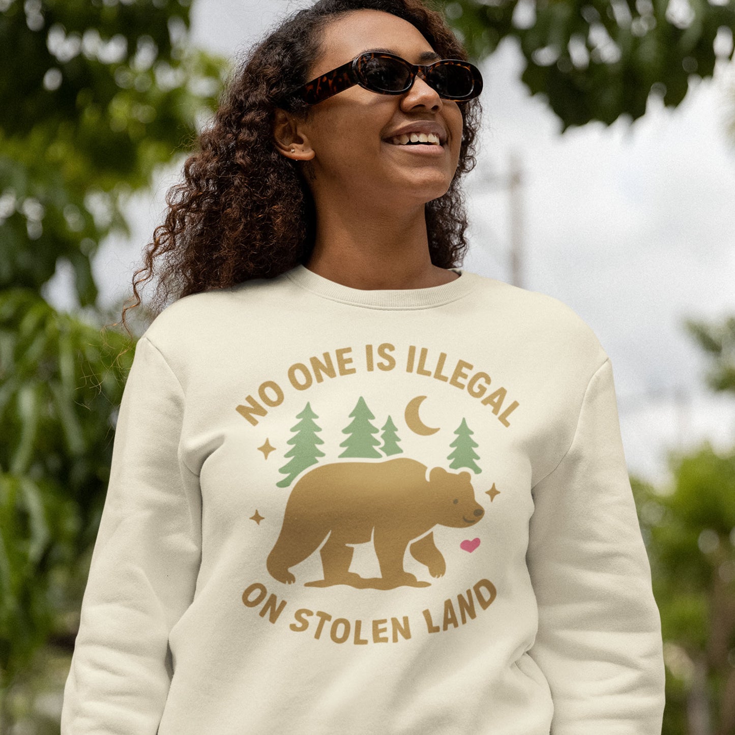Model wearing a cream unisex premium sweatshirt with the bear, pine trees, and moon illustration plus the message “NO ONE IS ILLEGAL ON STOLEN LAND,” styled for subtle Indigenous-rights advocacy.