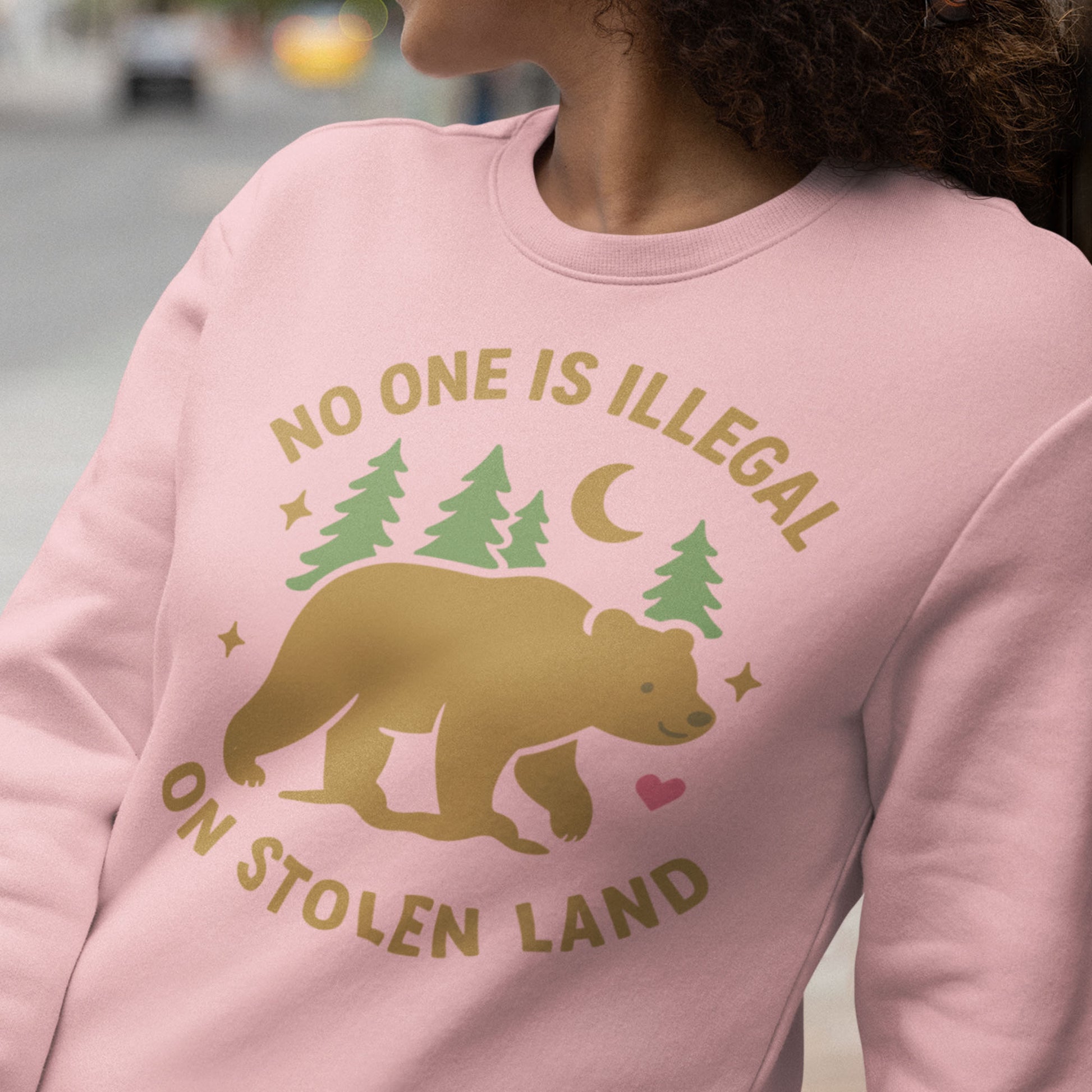 Model wearing a pink unisex premium sweatshirt displaying the bear, forest, and moon artwork with the message “NO ONE IS ILLEGAL ON STOLEN LAND,” a soft land-back inspired design from Blue Wave USA.