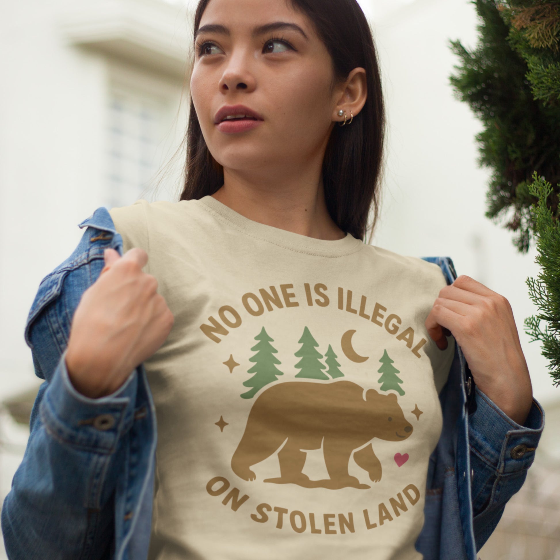 Model wearing a cream unisex t-shirt displaying the bear, forest, and moon graphic with the message “NO ONE IS ILLEGAL ON STOLEN LAND,” a soft activist tee from Blue Wave USA.