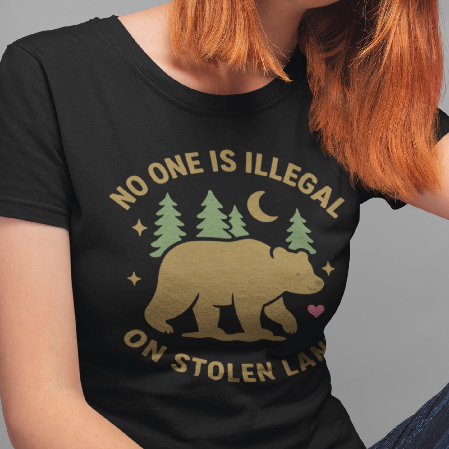 Model wearing a black women’s relaxed t-shirt showing the bear and forest design with the text “NO ONE IS ILLEGAL ON STOLEN LAND,” a soft activist tee from Blue Wave USA.