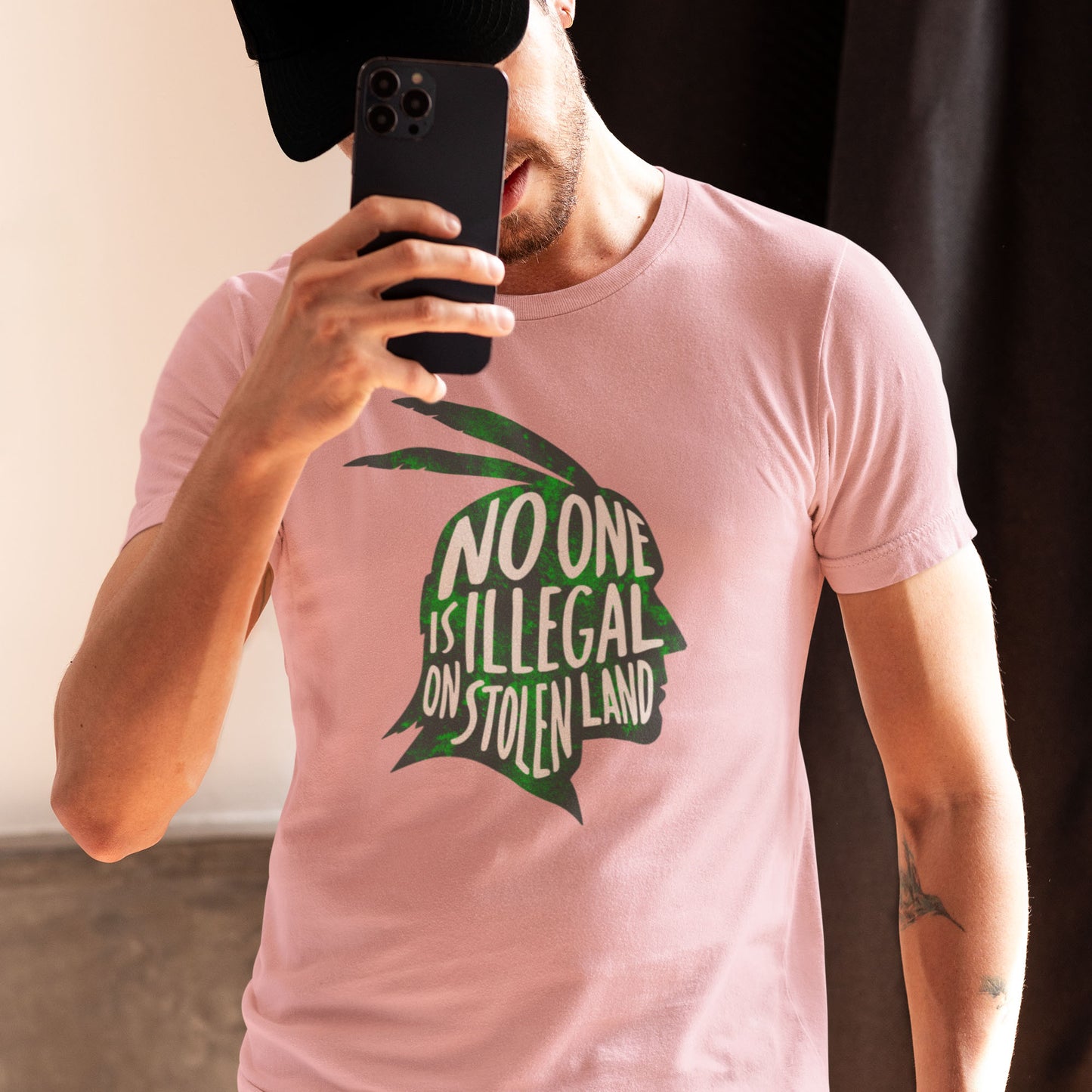 Person taking a mirror selfie wearing a pink unisex t-shirt displaying the green native american silhouette and message “NO ONE IS ILLEGAL ON STOLEN LAND,” offering a clean minimalist activist look.