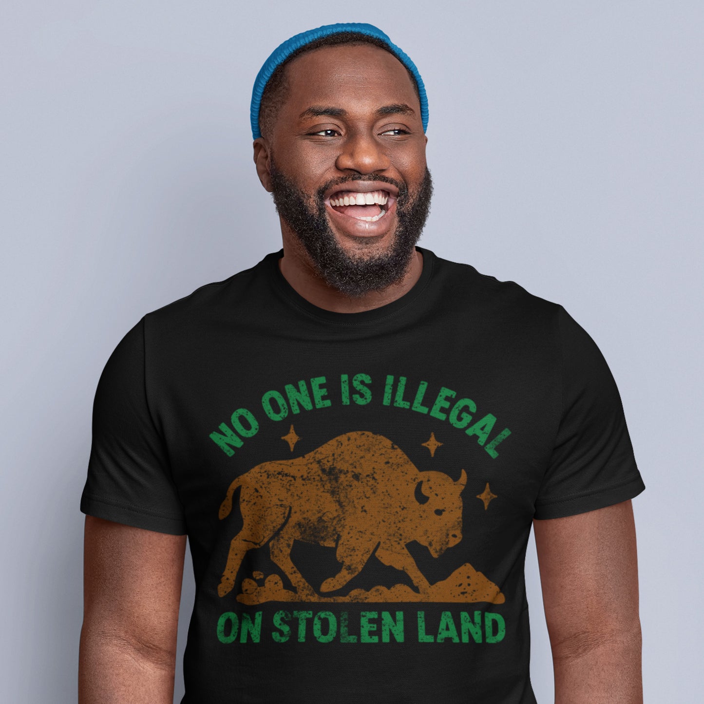 A smiling model wearing a black unisex tee printed with “NO ONE IS ILLEGAL ON STOLEN LAND” and a buffalo graphic, styled as a casual anti-colonial statement produced by Blue Wave USA.