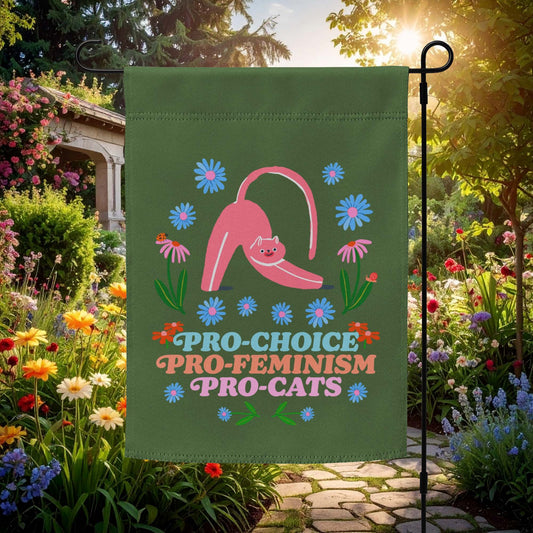 Green garden flag displayed outdoors in a flower garden, featuring a stretching pink cat surrounded by flowers and bold playful text reading “PRO-CHOICE PRO-FEMINISM PRO-CATS.”