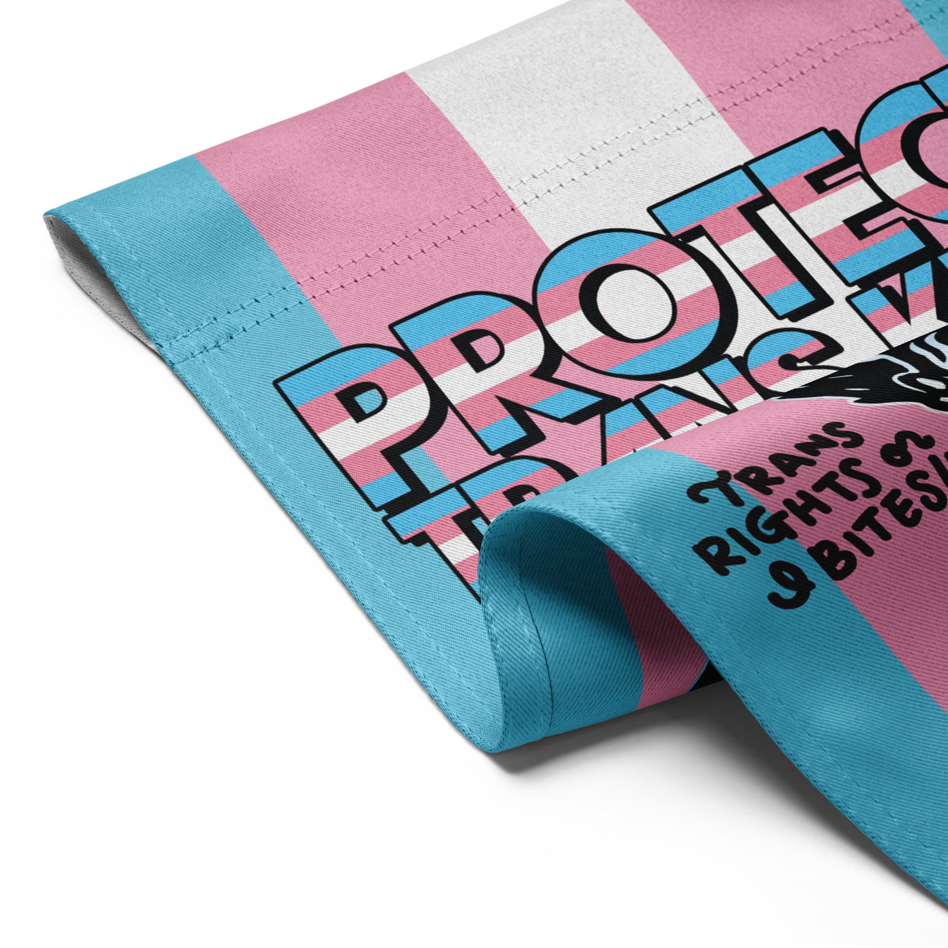 Folded garden flag revealing trans pride color bands, possum illustration details, and portions of the “Protect Trans Kids” message, highlighting fabric texture and print quality.