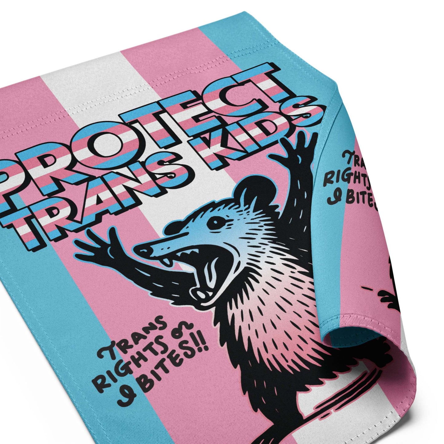 Close-up of garden flag fabric showing stitched edges, vibrant trans pride stripes, and detailed possum illustration beneath the “Protect Trans Kids” text.