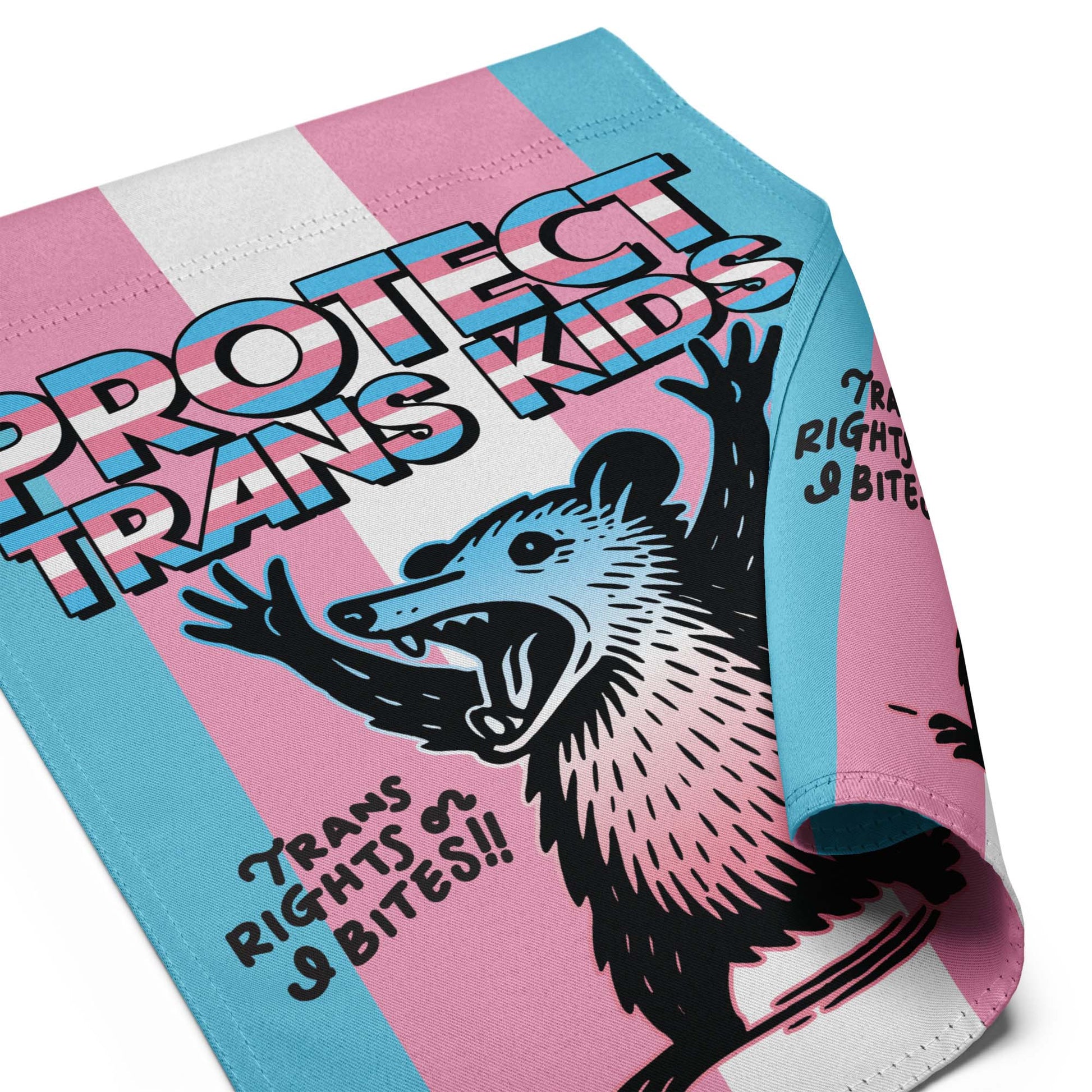 Close-up of garden flag fabric showing stitched edges, vibrant trans pride stripes, and detailed possum illustration beneath the “Protect Trans Kids” text.