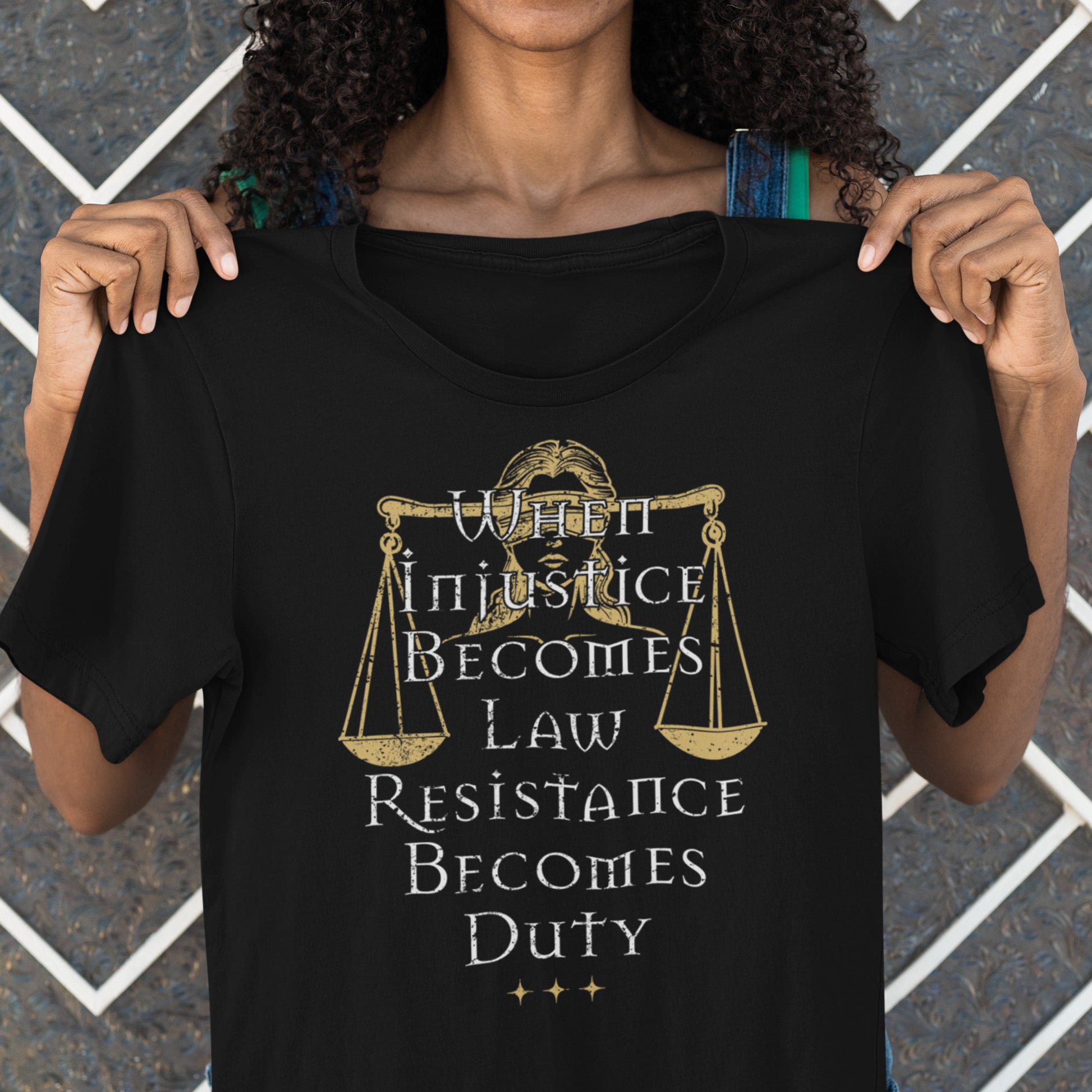 Model holding black unisex t-shirt featuring Lady Justice scales illustration with gold and white serif typography, centered progressive justice graphic from Blue Wave USA.