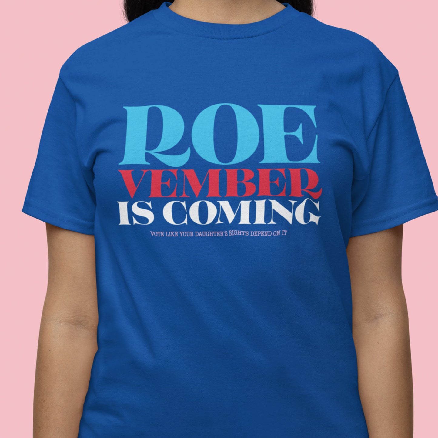 Model wearing royal blue unisex t-shirt featuring stacked typography reading “Roevember Is Coming” in patriotic colors.