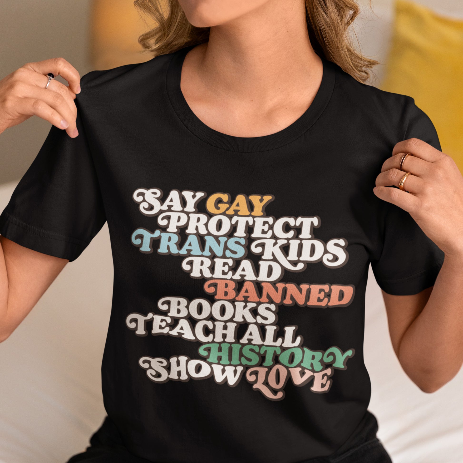 Model wearing black unisex t-shirt featuring retro rainbow-tone stacked typography reading “Say Gay Protect Trans Kids Read Banned Books Teach All History Show Love.”