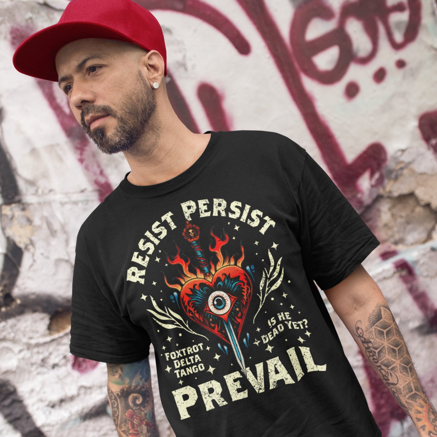 Model wearing black unisex t-shirt with flaming heart and dagger graphic, arched text “RESIST PERSIST,” bold “PREVAIL,” and small phrases “FOXTROT DELTA TANGO” and “IS HE DEAD YET?”