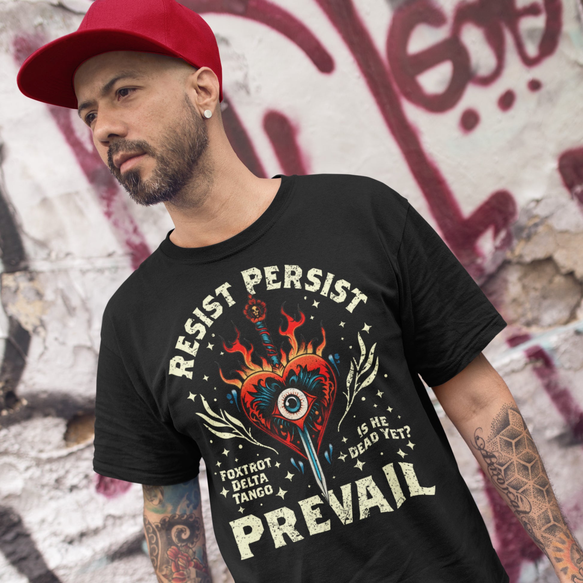 Model wearing black unisex t-shirt with flaming heart and dagger graphic, arched text “RESIST PERSIST,” bold “PREVAIL,” and small phrases “FOXTROT DELTA TANGO” and “IS HE DEAD YET?”