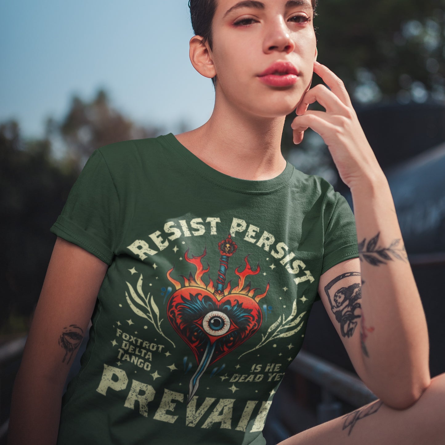 Model wearing forest green unisex t-shirt with flaming heart and dagger graphic, distressed arched text “RESIST PERSIST,” bold “PREVAIL,” and small text “FOXTROT DELTA TANGO.”