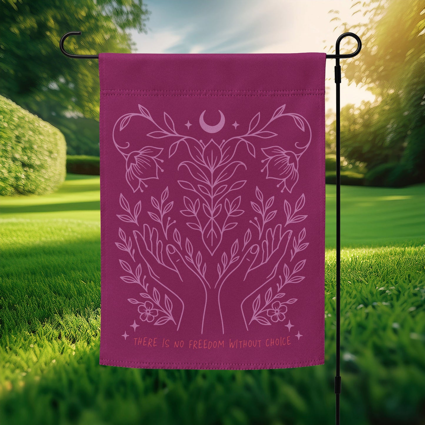 Pro-choice garden flag displayed in sunlight, featuring a pink line art design of hands, leaves, and a crescent moon on a deep plum background with the phrase “There Is No Freedom Without Choice.”