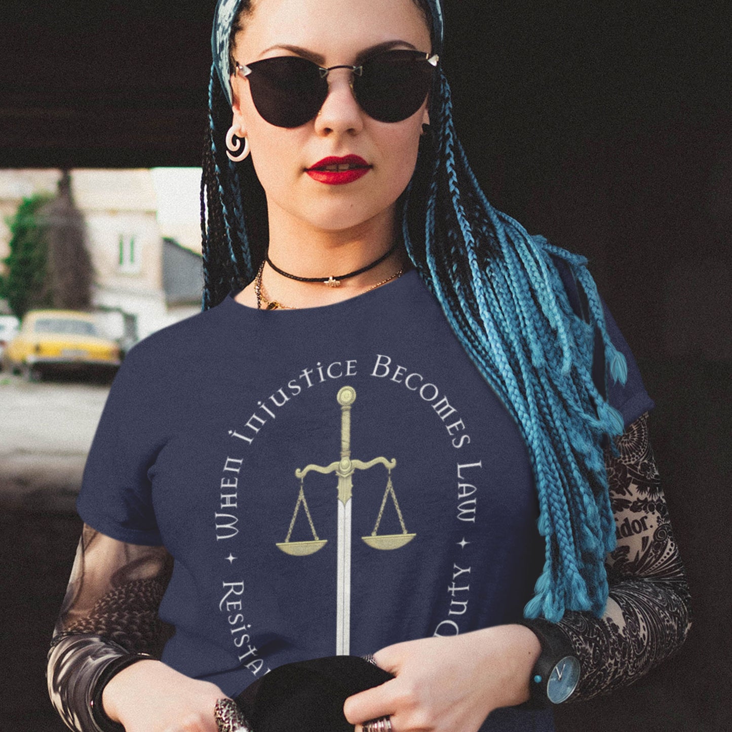 Person with blue braids and sunglasses wearing the navy “When Injustice Becomes Law” t-shirt. The design’s gold scale glows against the dark fabric — a Blue Wave USA call for courage and civil resistance.