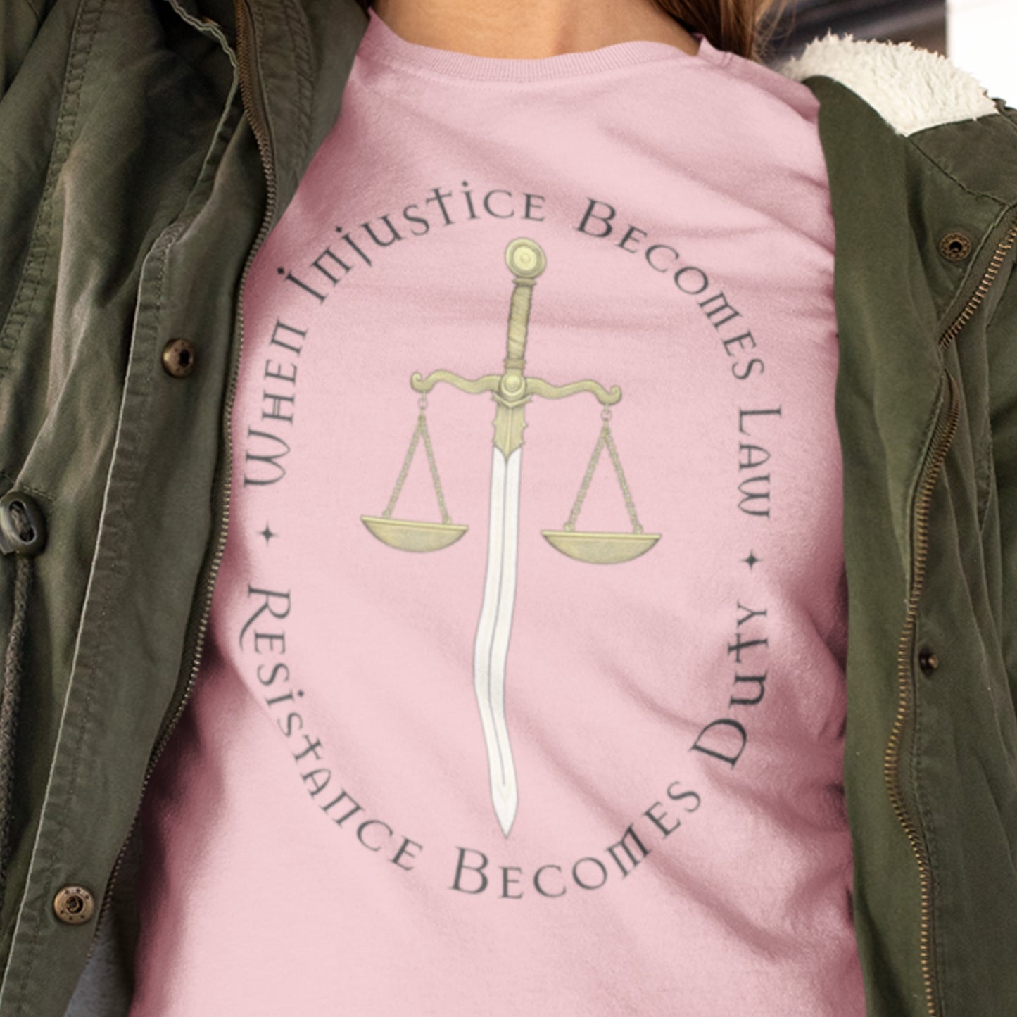 Close-up of a person wearing a pink version of the “When Injustice Becomes Law” shirt, highlighting the gold justice scale and fine serif typography.