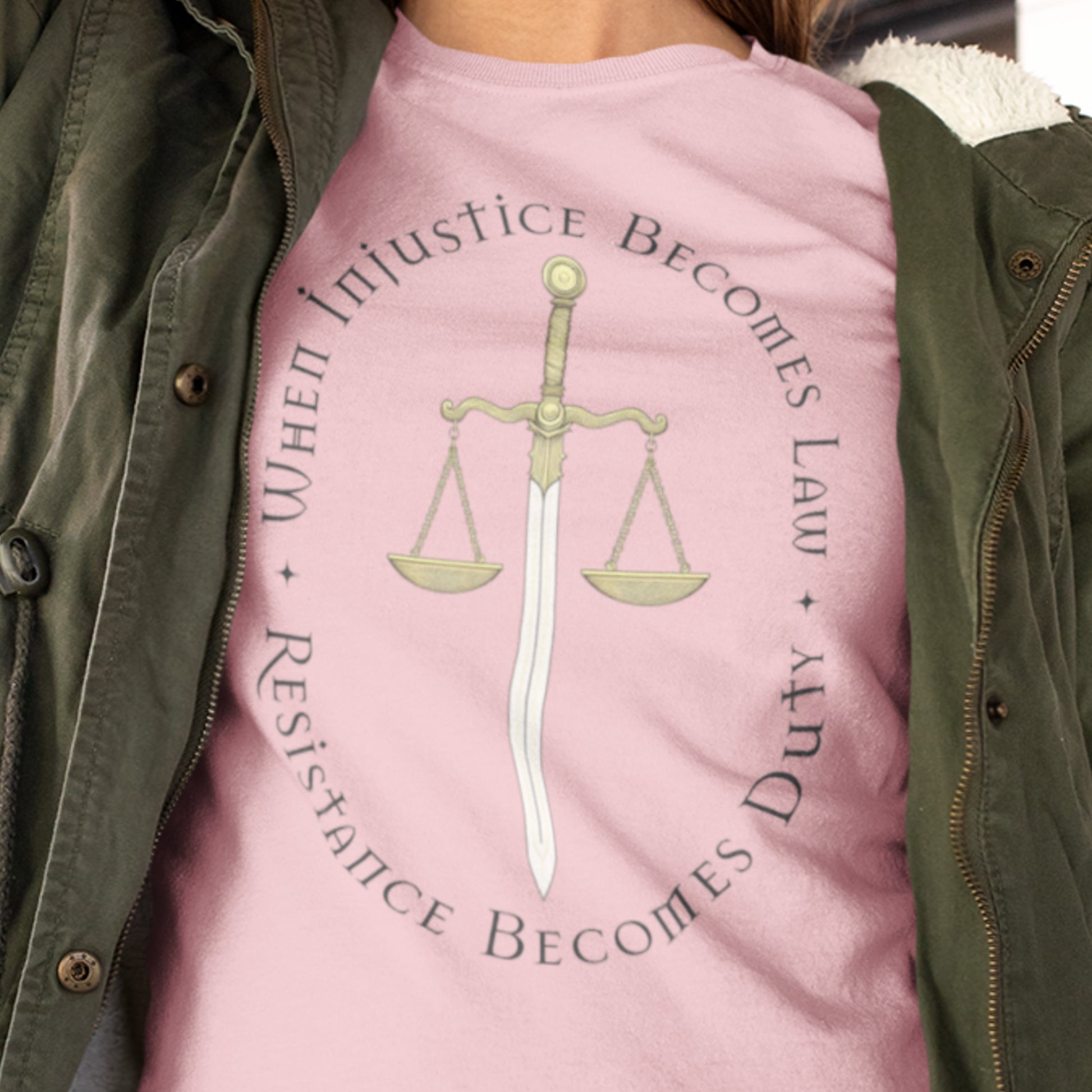 Close-up of a person wearing a pink version of the “When Injustice Becomes Law” shirt, highlighting the gold justice scale and fine serif typography.
