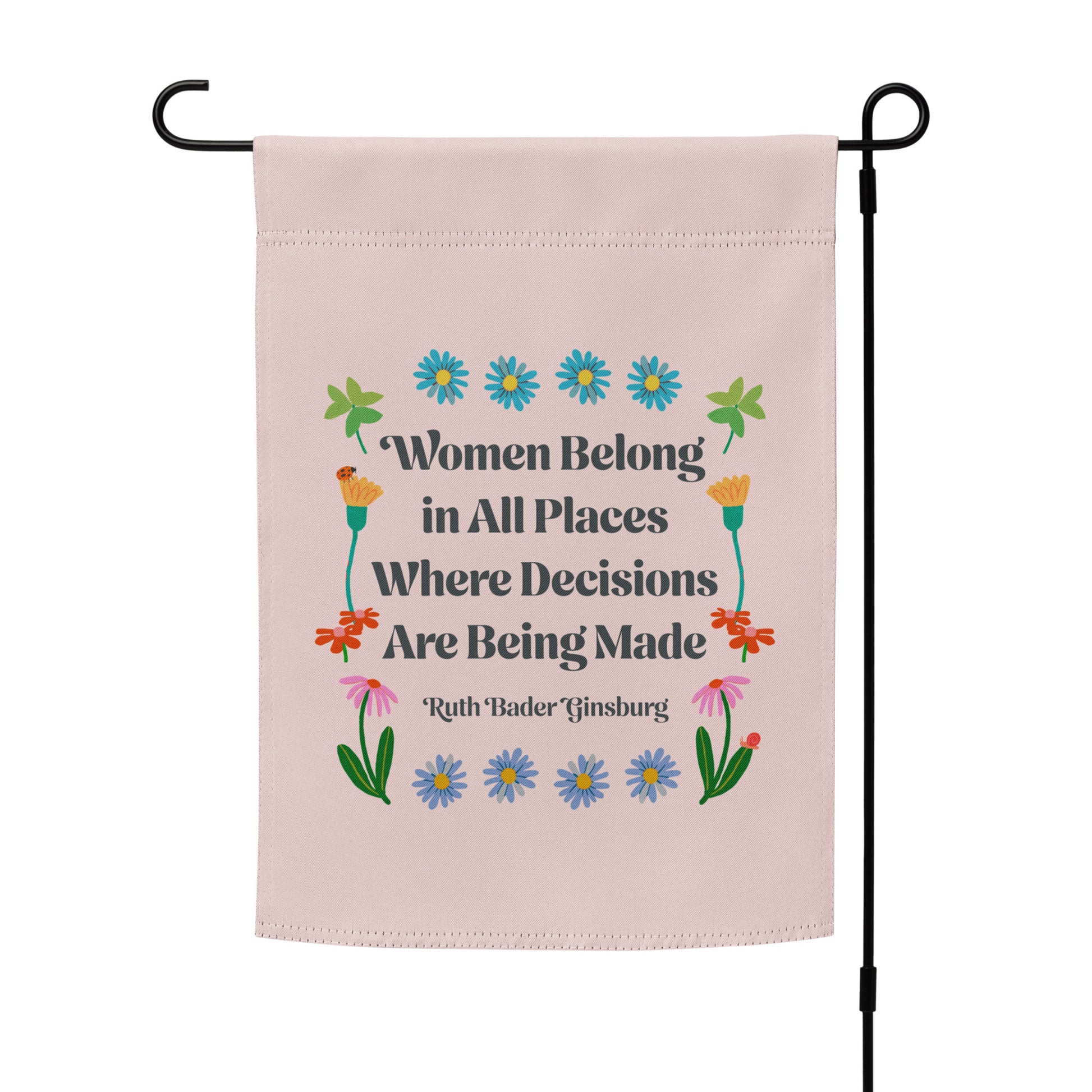 Light pink garden flag displayed vertically on a black metal stand, featuring floral motifs and centered text reading “Women Belong in All Places Where Decisions Are Being Made.”