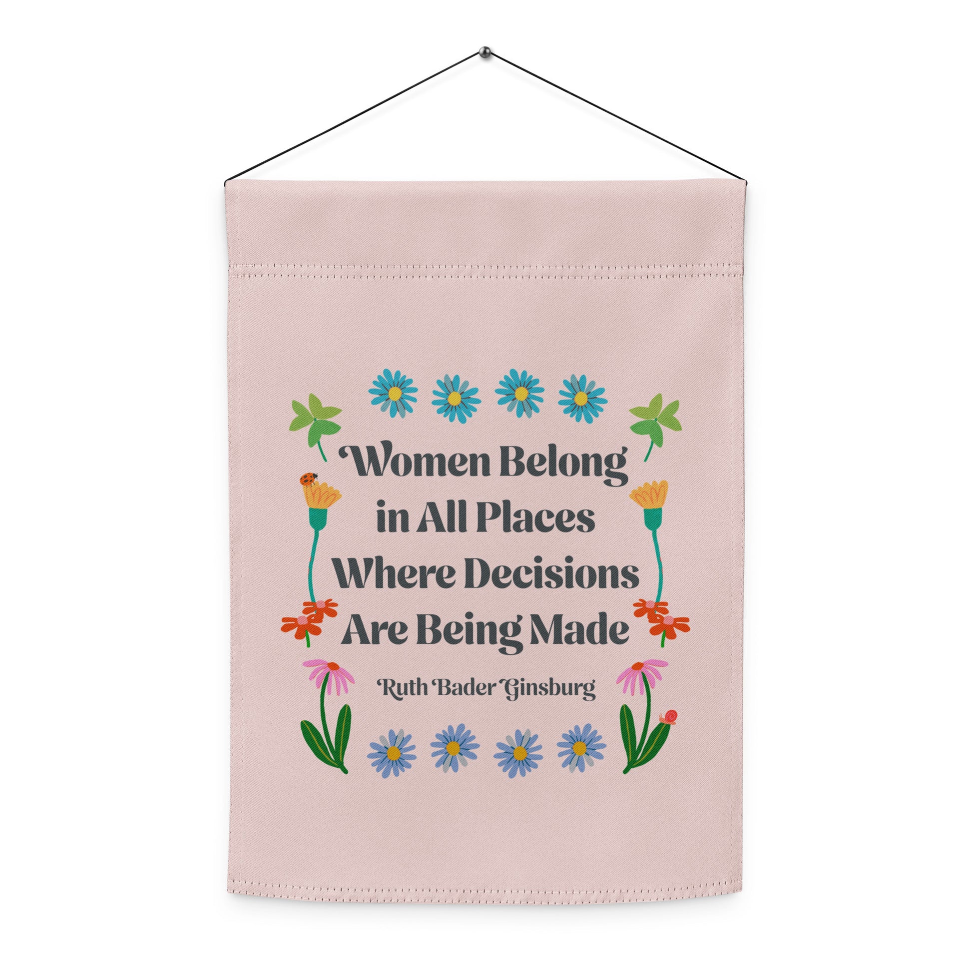Indoor hanging view of the light pink garden flag, showcasing the full floral folk-art layout and the RBG quote “Women Belong in All Places Where Decisions Are Being Made,” by Blue Wave USA.
