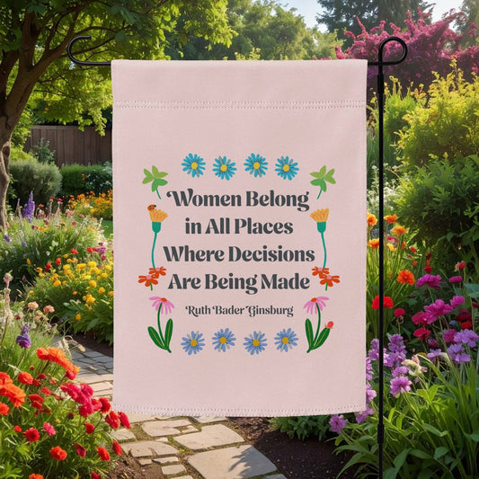 Light pink garden flag displayed outdoors in a flower garden, featuring floral folk-art illustrations and serif typography reading “Women Belong in All Places Where Decisions Are Being Made.”