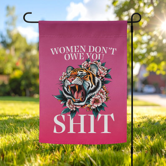 Deep pink garden flag displayed outdoors on a metal stand, featuring a roaring tiger illustration with floral accents and bold serif text reading “WOMEN DON’T OWE YOU SH*T.”