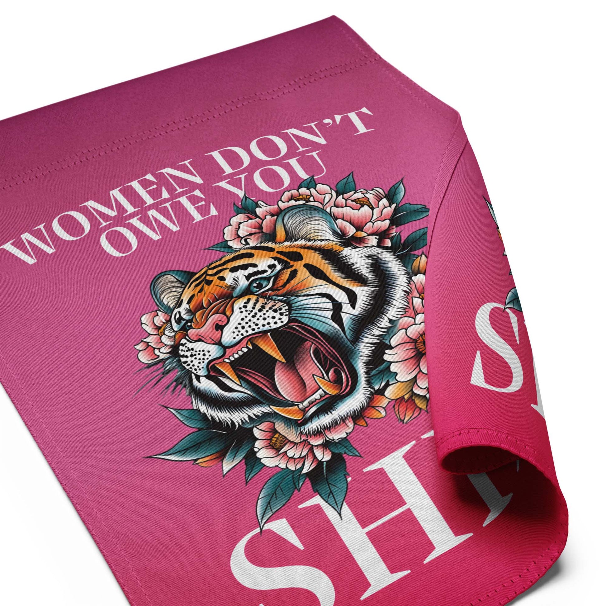 Folded pink garden flag shown close up, highlighting the smooth fabric texture, stitched edges, and partial view of the tiger illustration and text reading “WOMEN DON’T OWE YOU SH*T.”
