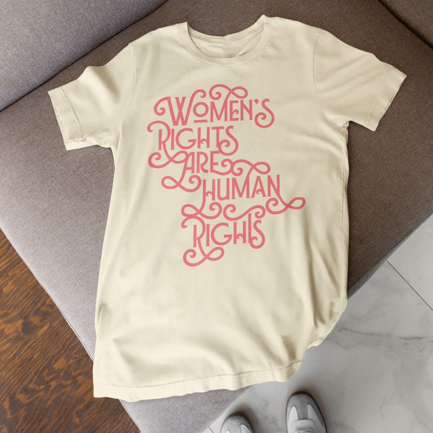 Cream “Women’s Rights Are Human Rights” shirt folded neatly on a bed with minimalist décor, showing delicate coral lettering.