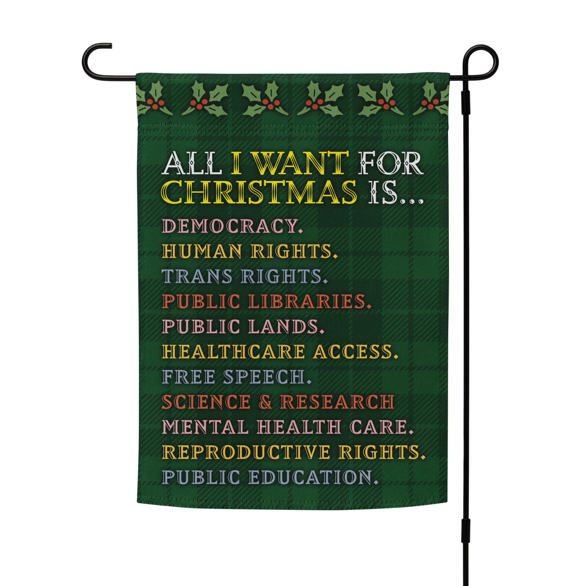 Green plaid garden flag on a metal stand featuring holly motifs and the printed message “ALL I WANT FOR CHRISTMAS IS…” followed by items such as human rights and free speech, designed by Blue Wave USA.