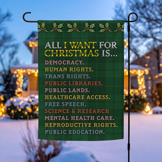 Green garden flag on a metal stand with plaid background, holly border, and the printed list beginning “ALL I WANT FOR CHRISTMAS IS…” including democracy and human rights; progressive décor by Blue Wave USA.