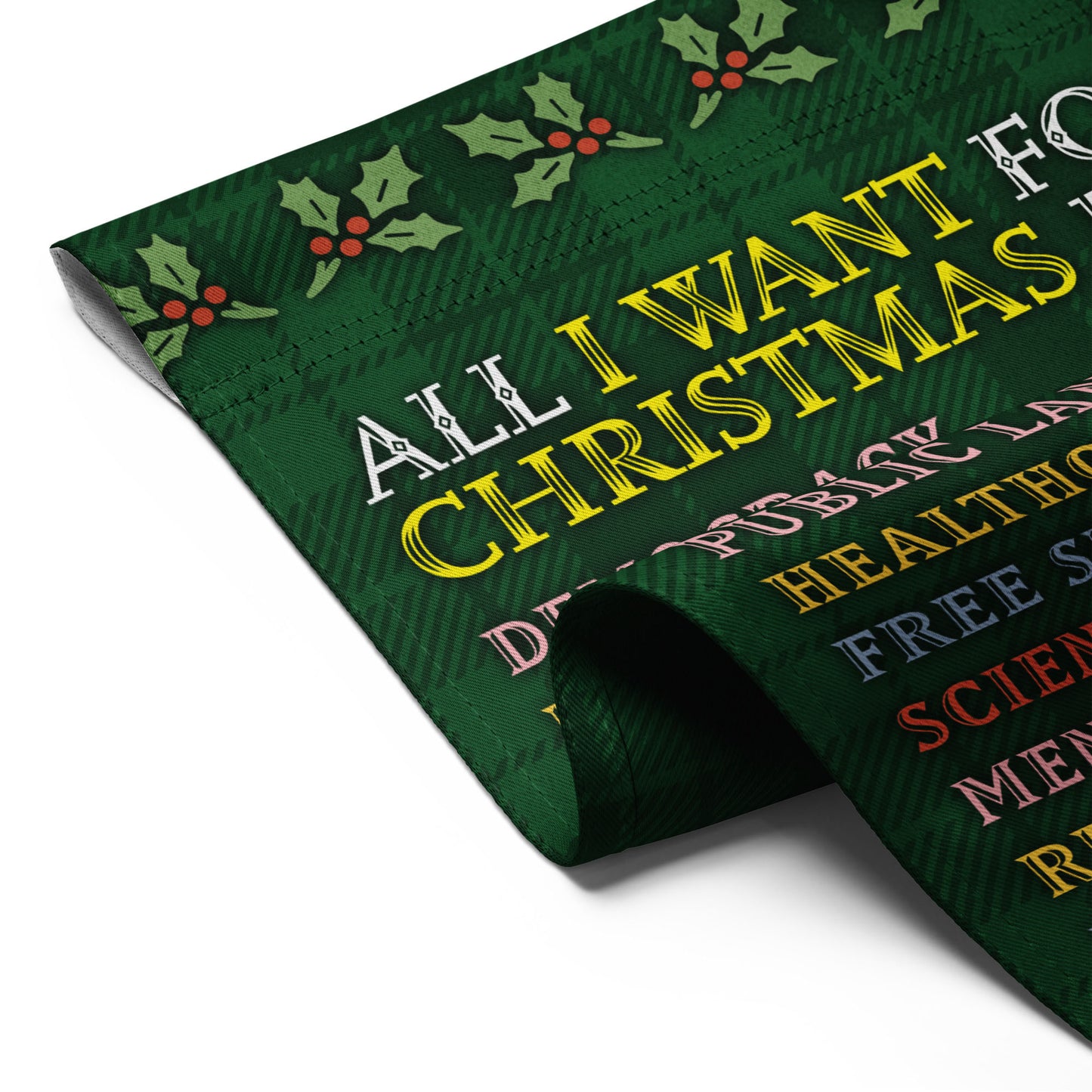 Close-up of folded green garden flag fabric showing plaid texture, holly design, and partial text from the “ALL I WANT FOR CHRISTMAS IS…” progressive holiday list.