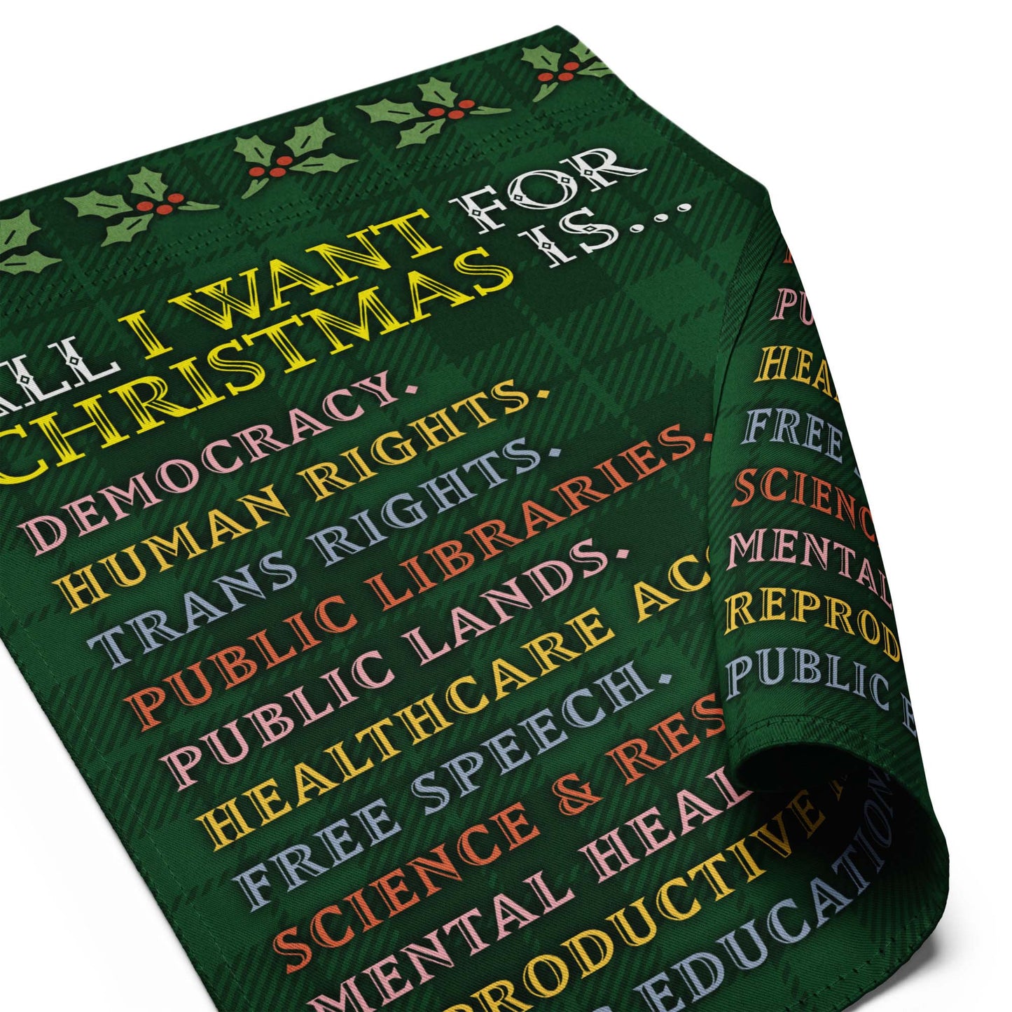 Rolled green garden flag displaying plaid background, holly border, and partial bold text from the “ALL I WANT FOR CHRISTMAS IS…” printed list.