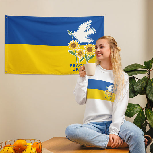 Person sitting with coffee in front of a hanging “Peace for Ukraine” flag featuring dove with olive branch and sunflowers on blue and yellow background, sold by Blue Wave USA progressive products.