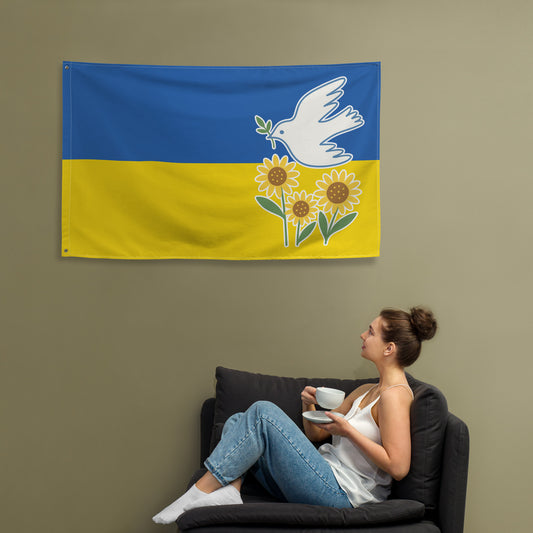 Person sitting with coffee in front of a hanging “Ukraine Peace and Solidarity” flag featuring white dove with olive branch and sunflowers on a blue and yellow background, sold by Blue Wave USA progressive products.