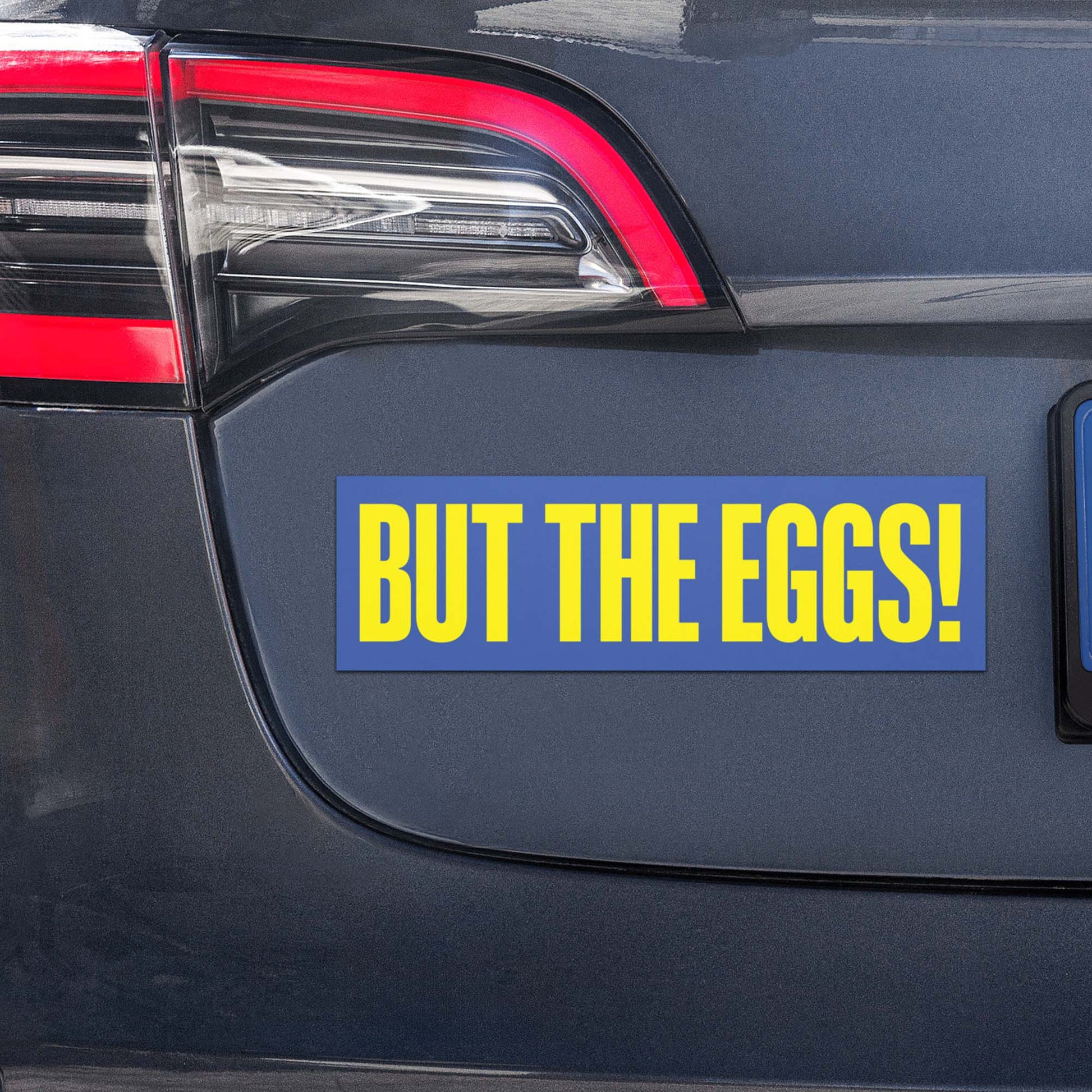 Anti-Trump Satire car bumper magnet displayed on vehicle bumper with bold yellow text reading “BUT THE EGGS!” on blue background.