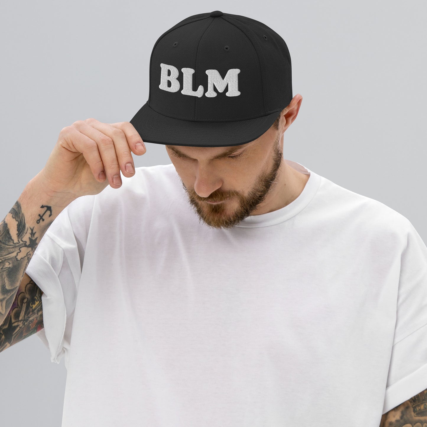 Model adjusting a black snapback hat featuring bold embroidered “BLM” text, structured crown and flat brim, shown in a neutral studio setting.