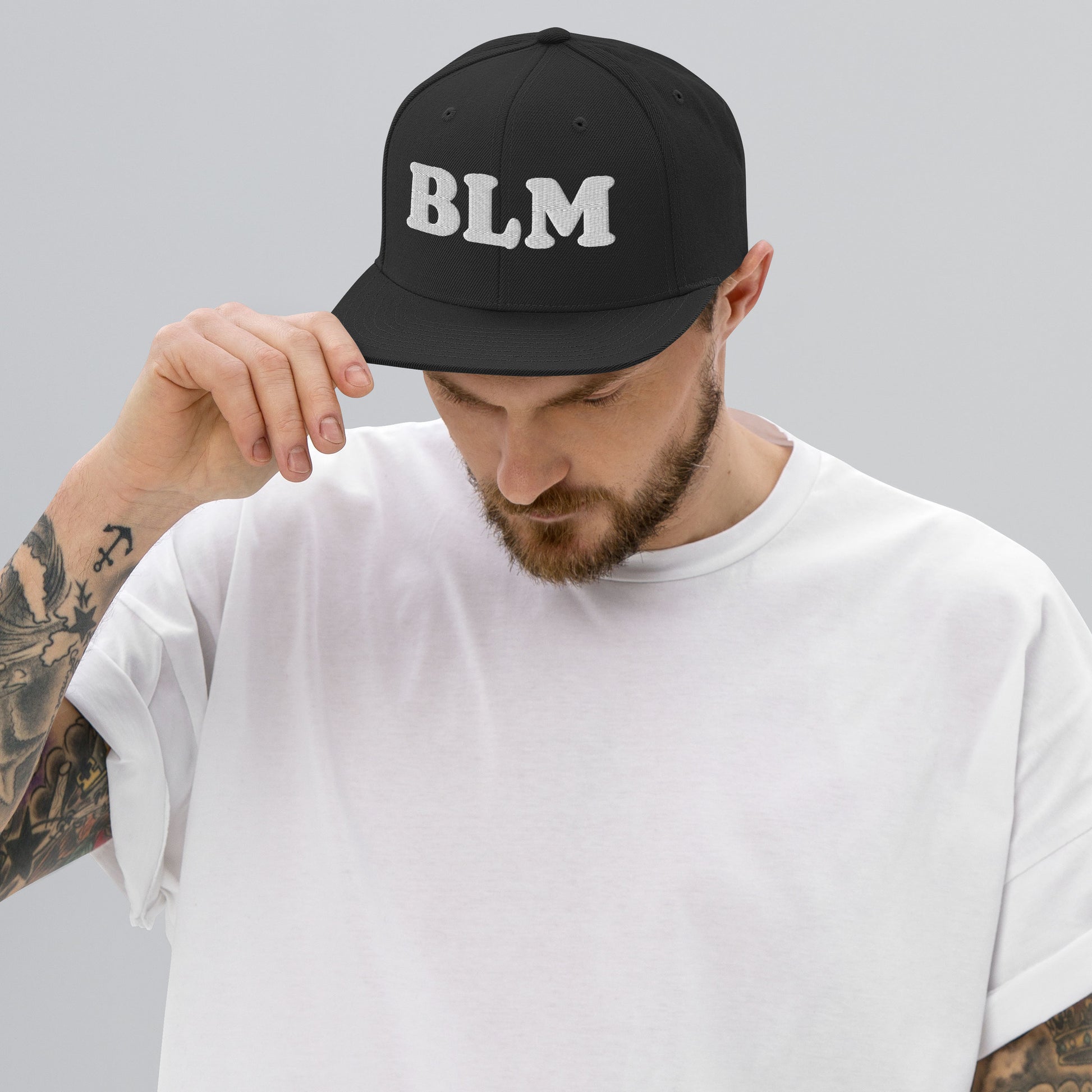 Model adjusting a black snapback hat featuring bold embroidered “BLM” text, structured crown and flat brim, shown in a neutral studio setting.