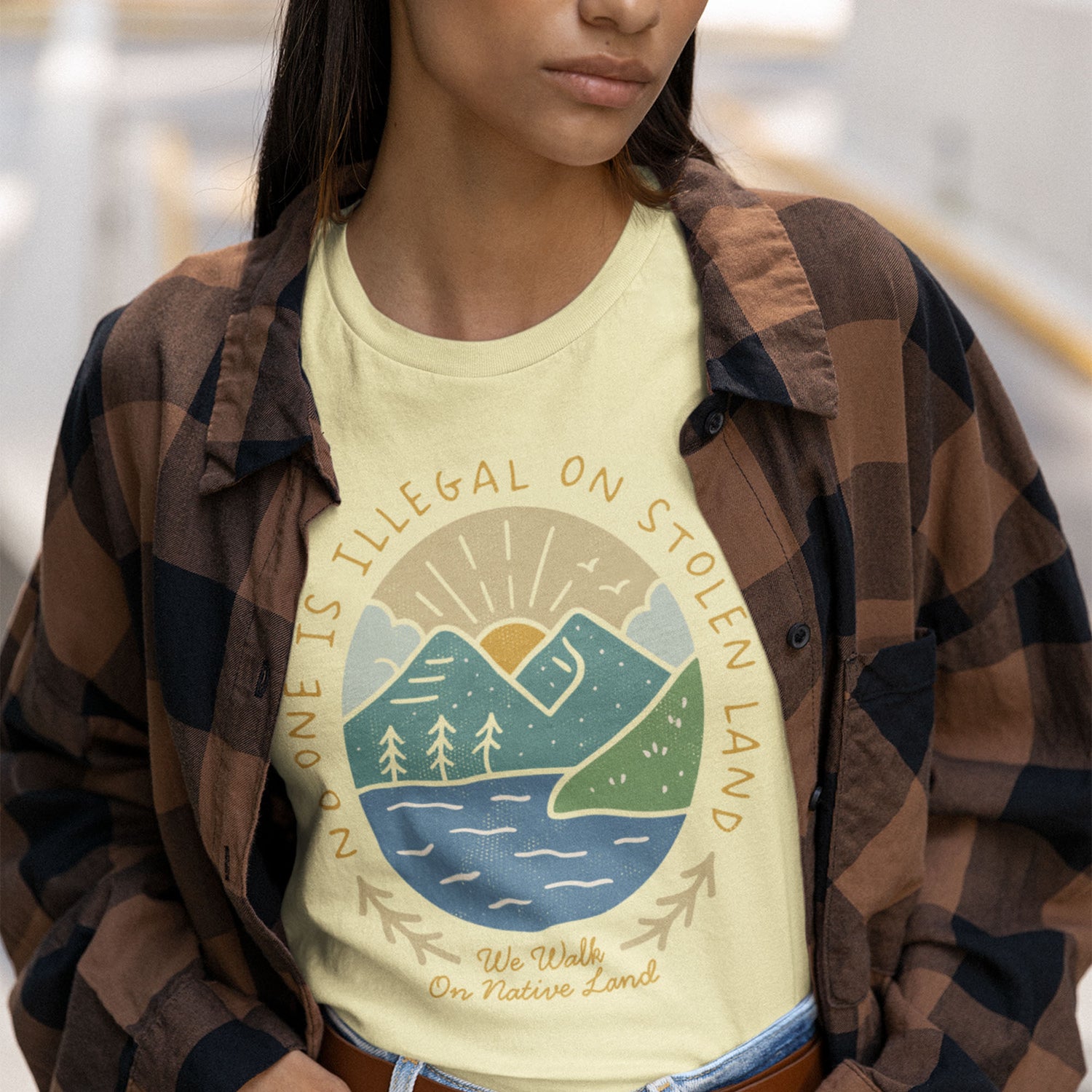 Person wearing the cream “No One Is Illegal on Stolen Land” tee layered under a brown plaid shirt, styled for a casual activist look.