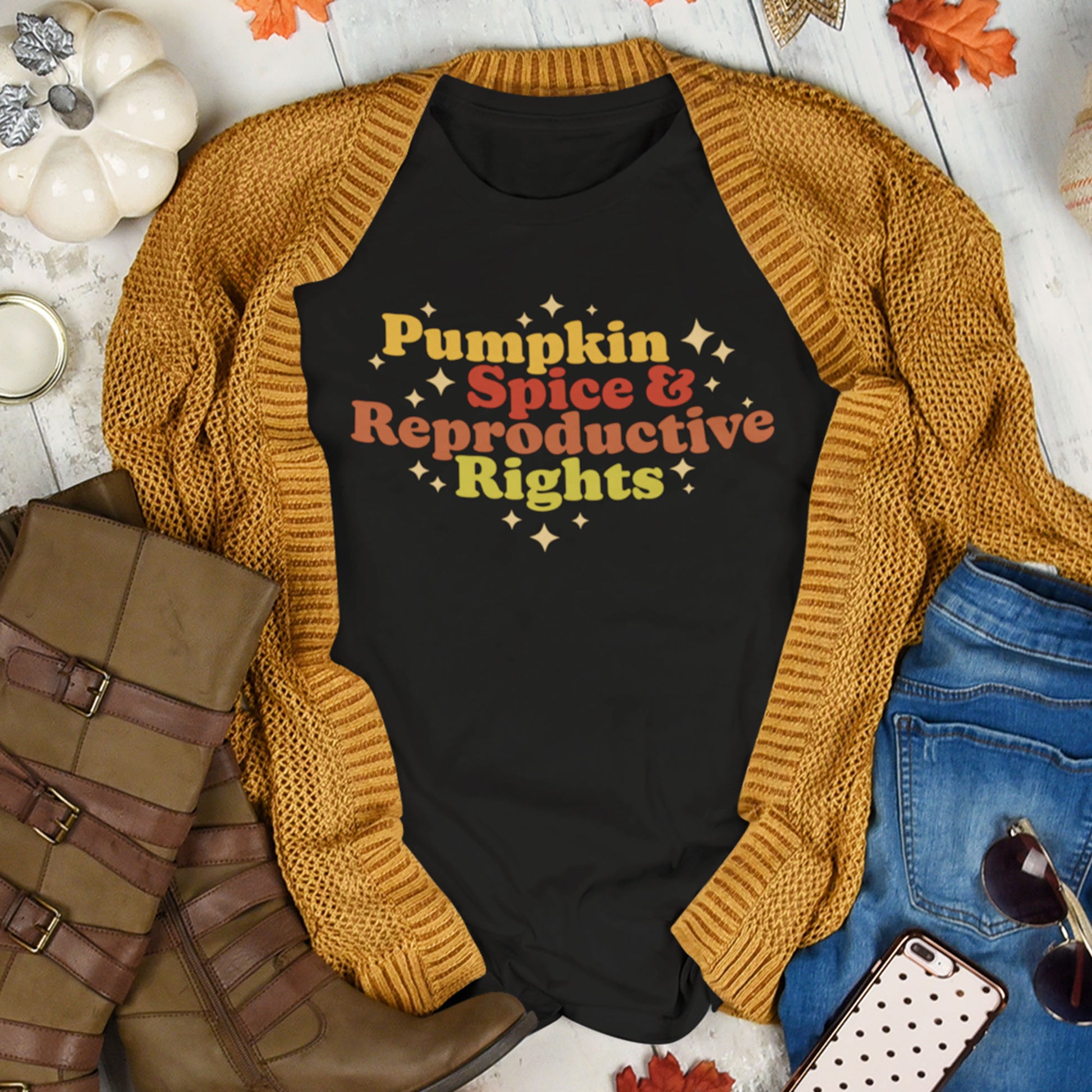 Black t-shirt with 'Pumpkin Spice & Reproductive Rights' text, mustard sweater, jeans, boots, and phone on a wooden surface.