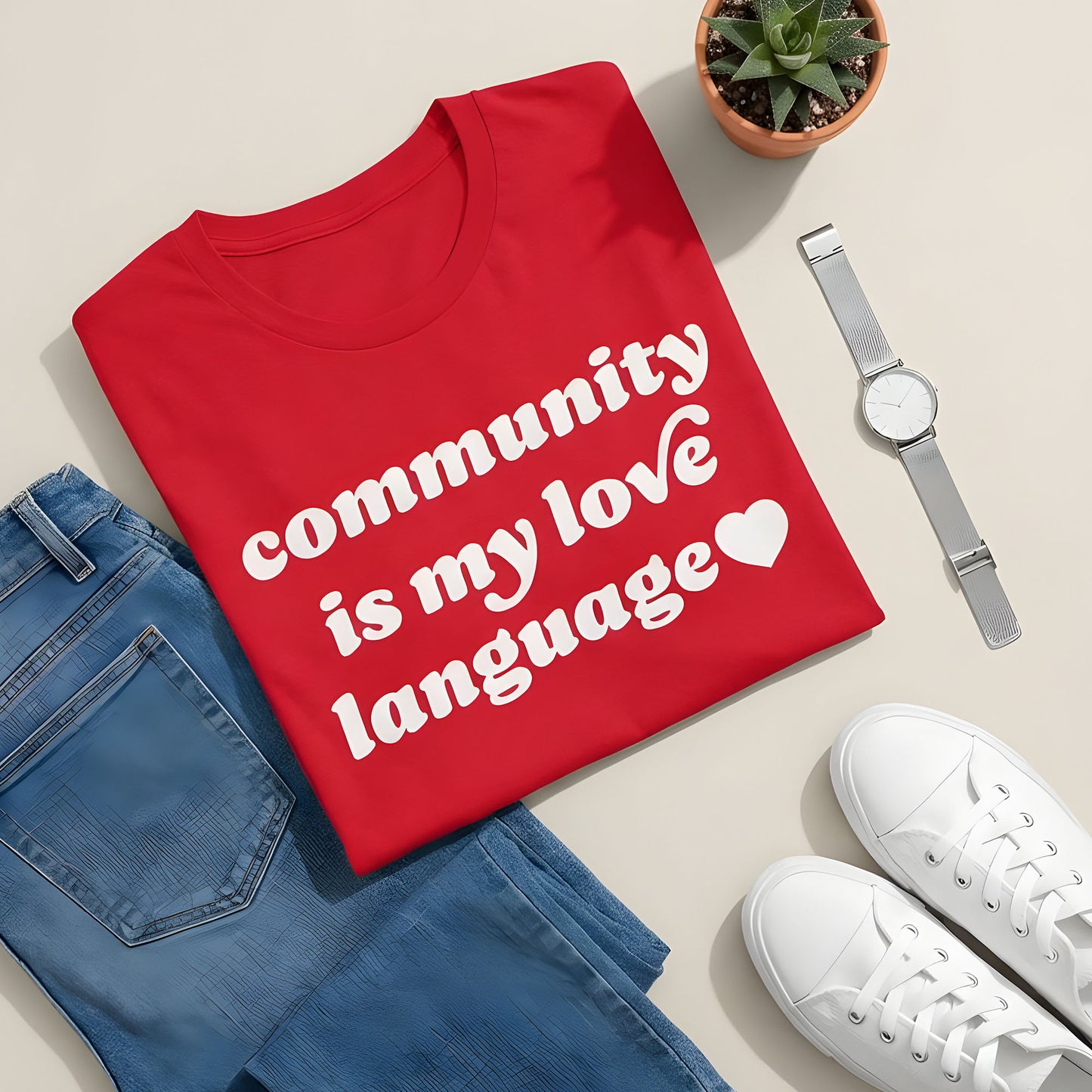 Red unisex t-shirt in a styled flatlay with jeans, sneakers, watch, and plant, featuring bold white retro lettering and heart icon reading “community is my love language.”