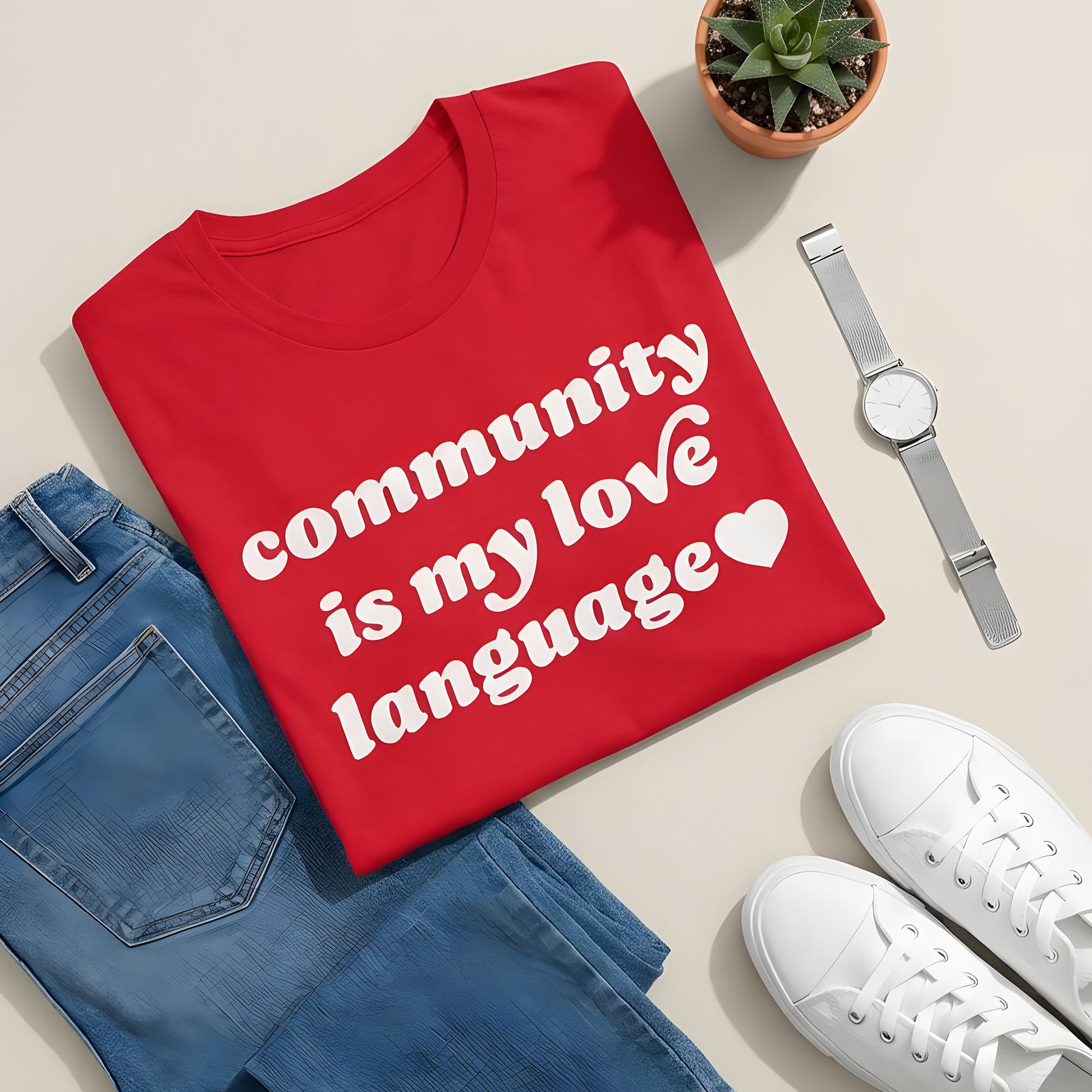 Red unisex t-shirt in a styled flatlay with jeans, sneakers, watch, and plant, featuring bold white retro lettering and heart icon reading “community is my love language.”