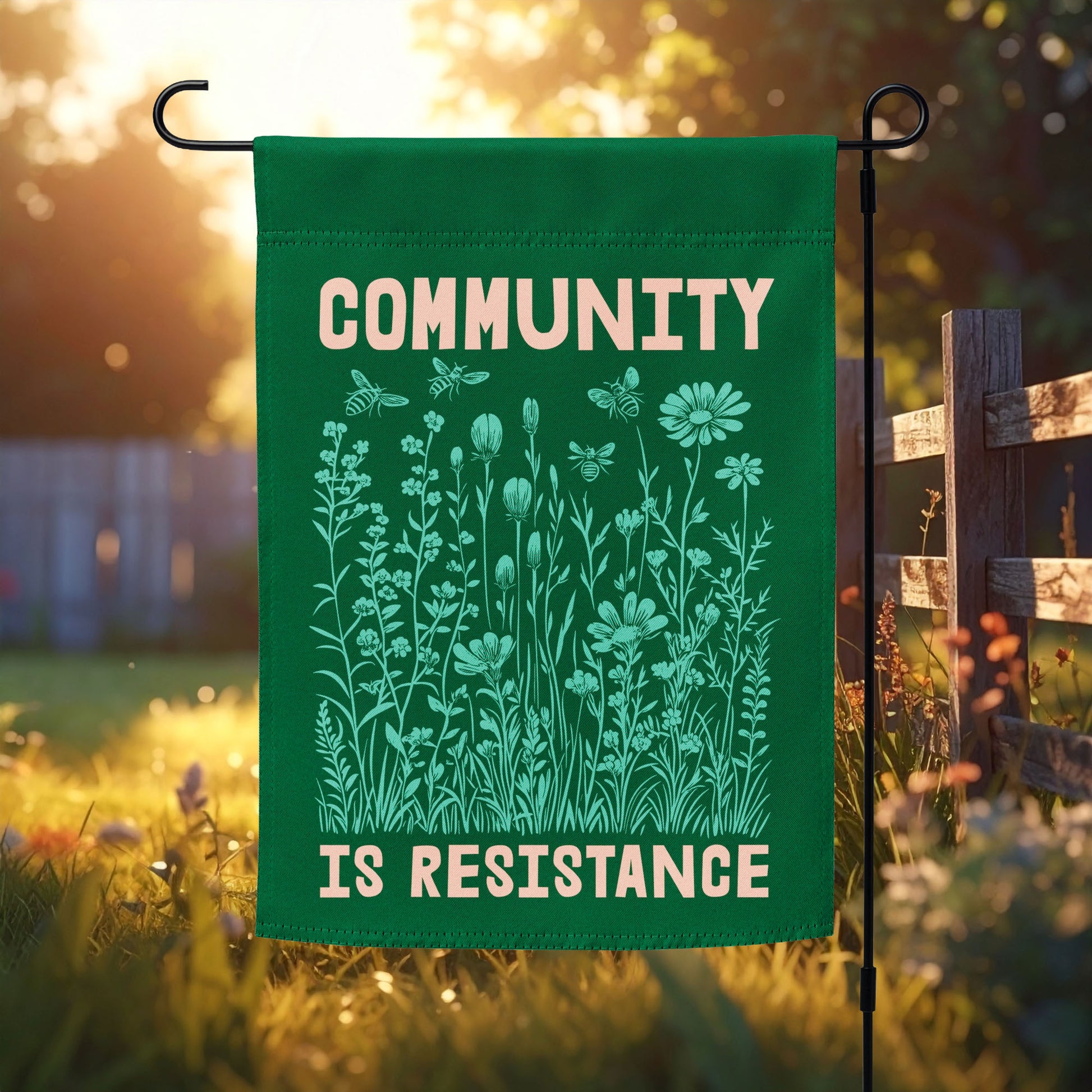 Green garden flag featuring mint-green wildflowers and soft cream text reading “COMMUNITY IS RESISTANCE,” displayed in morning sunlight beside a wooden fence. A hopeful outdoor statement for solidarity and care.