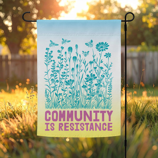Pastel garden flag featuring mint-blue wildflowers and magenta text reading “COMMUNITY IS RESISTANCE.” The flag glows softly in the morning sun, symbolizing renewal, hope, and collective care.