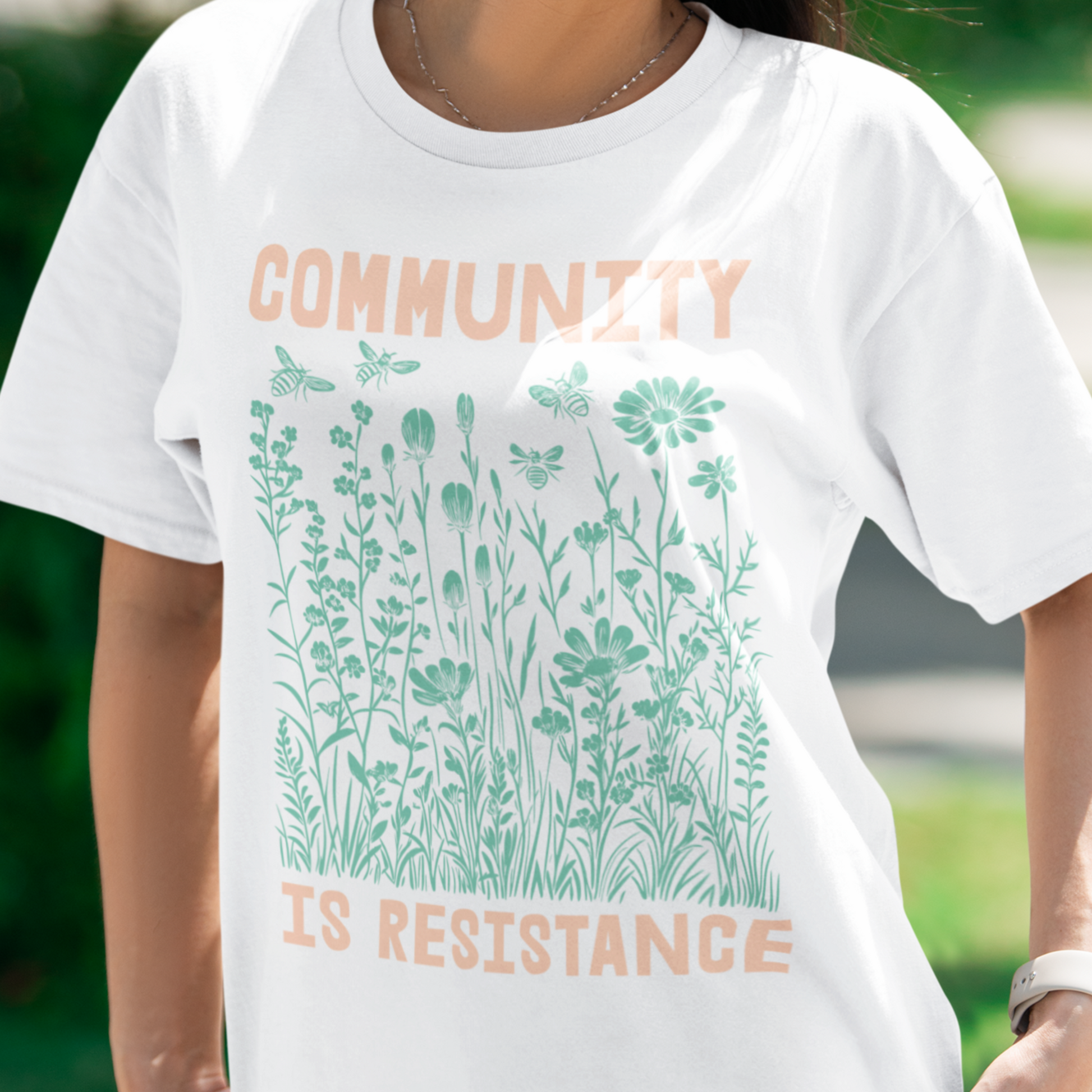 Person wearing a white Community Is Resistance t-shirt, standing outside in natural light. The pastel print of blooming wildflowers feels calm yet defiant — a quiet act of solidarity designed by Blue Wave USA.