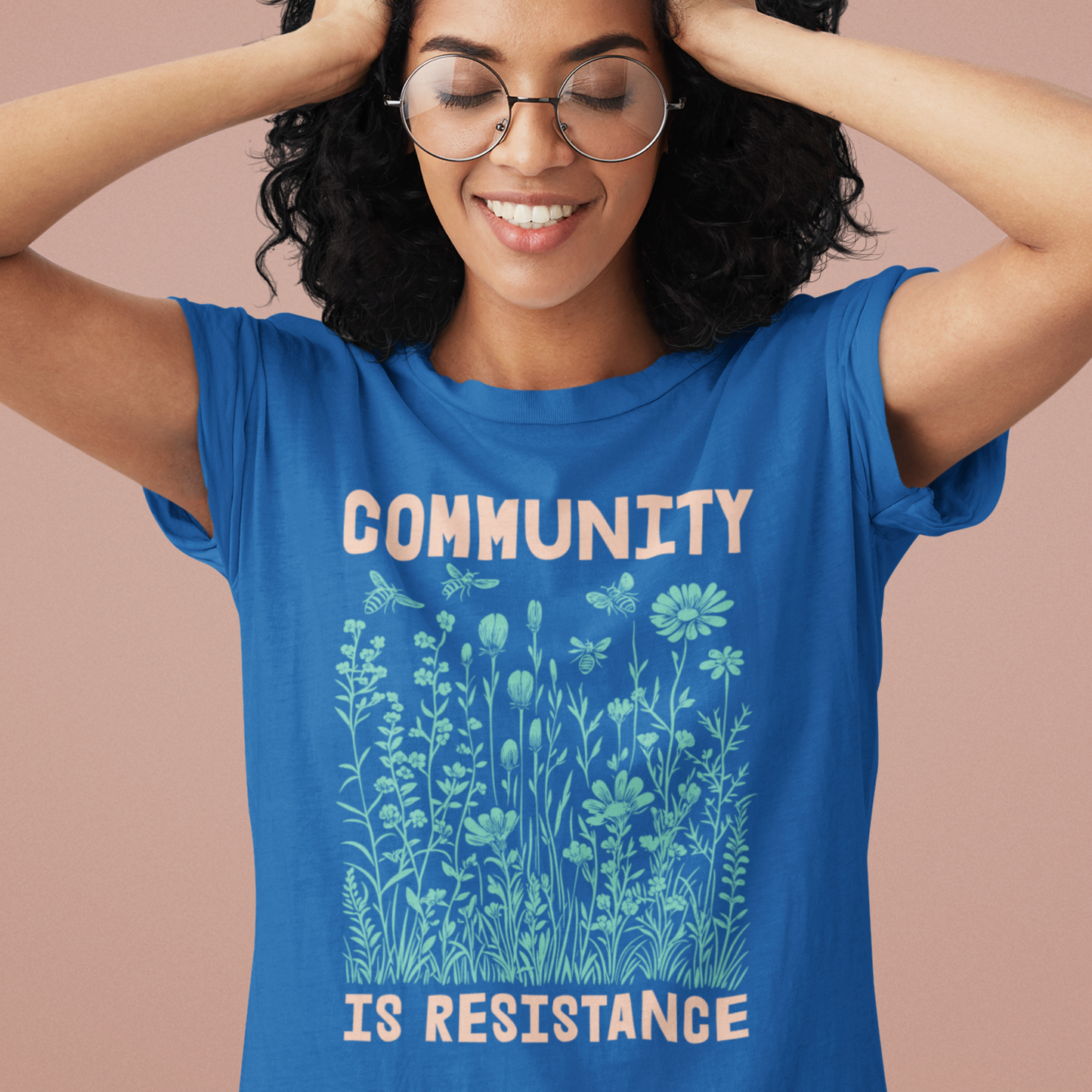 Person smiling with hands raised, wearing a royal blue Community Is Resistance tee. The soft green botanical pattern across the chest celebrates grassroots strength and interconnected resistance.