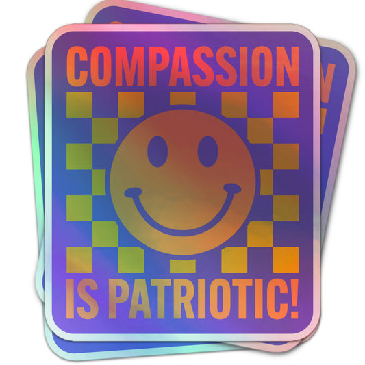 Holographic sticker with a large smiley face, yellow checkerboard pattern, and bold text reading “Compassion is Patriotic!”