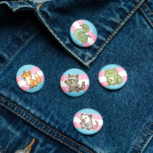 Five trans pride pin buttons attached to a denim jacket, showcasing cute animal artwork and transgender flag-inspired pink, blue, and white backgrounds.