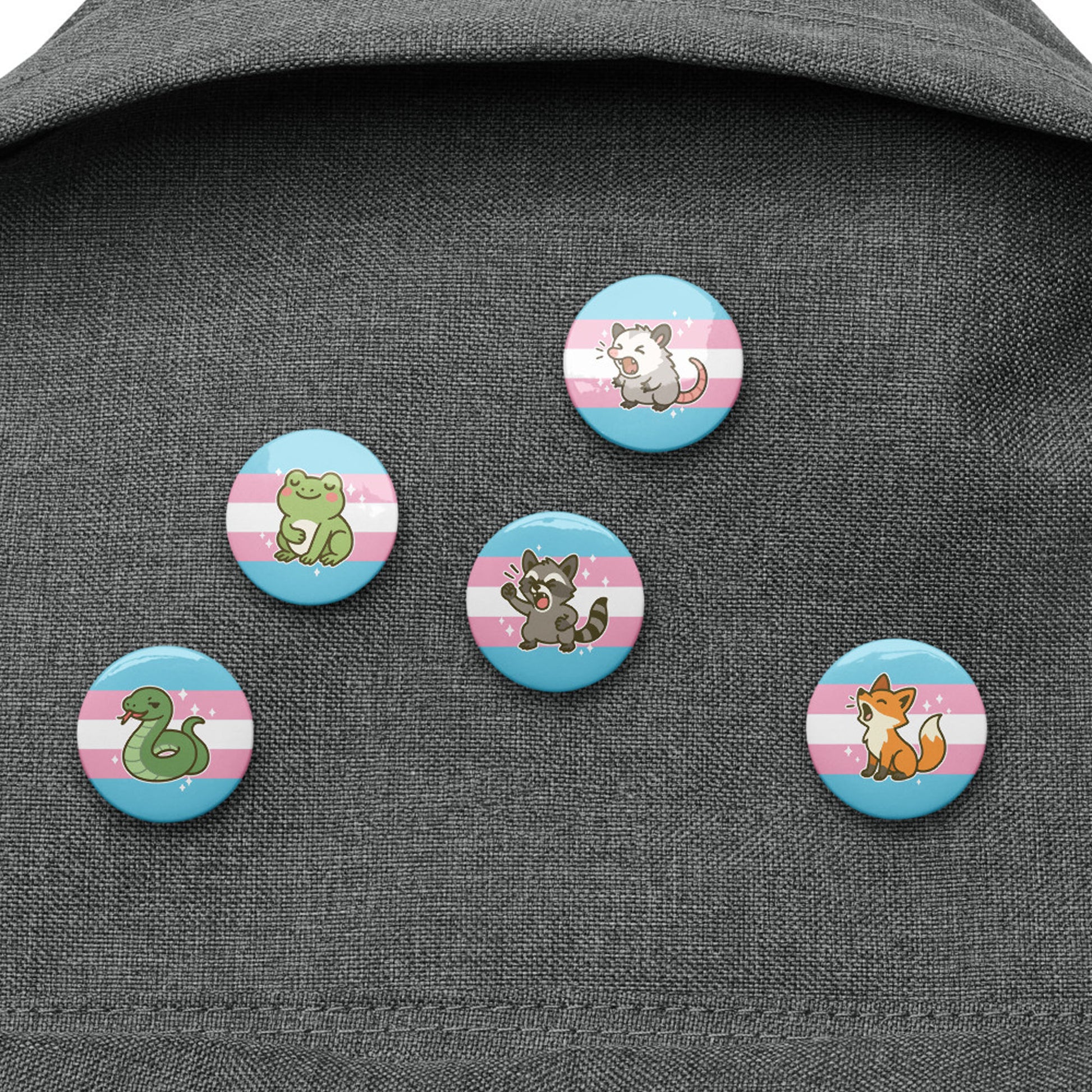 Trans rights pin button collection displayed on dark fabric backpack, featuring five adorable animal designs inspired by possums, frogs, foxes, raccoons, and snakes with trans pride colors.