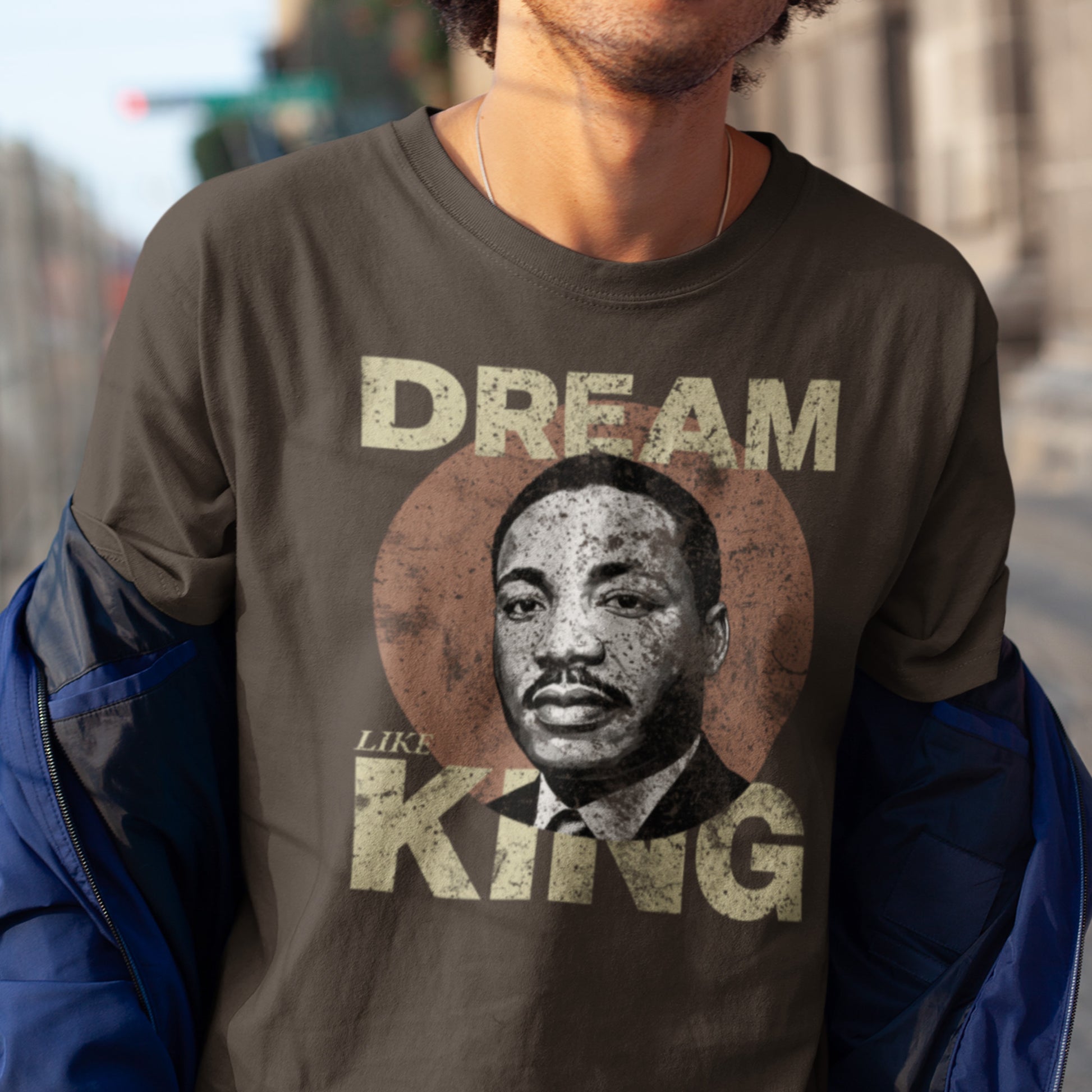 Man wearing a brown unisex t-shirt featuring the “DREAM LIKE KING” graphic with MLK’s portrait inside a rustic circle. Vintage civil-rights look for casual activism.