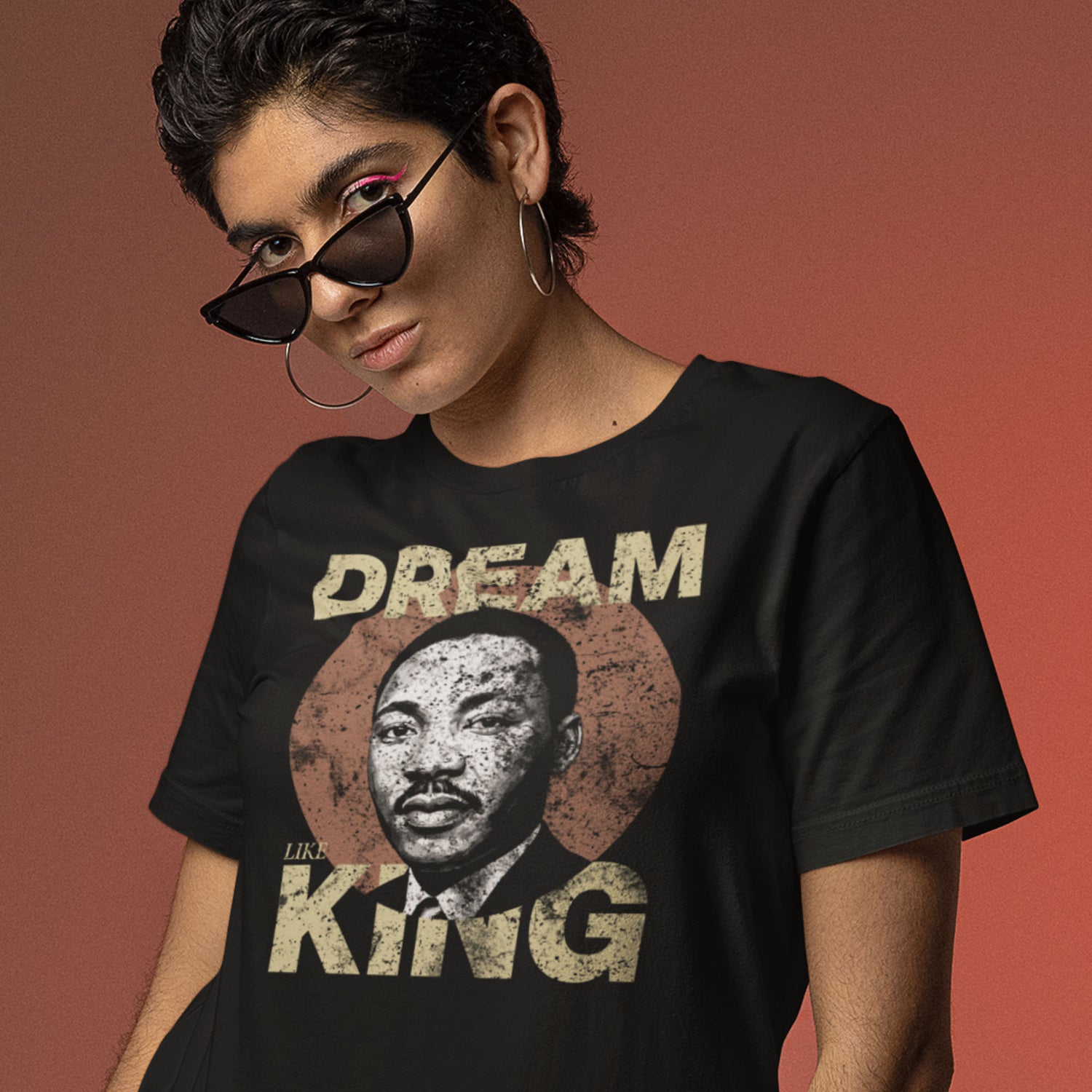 Woman wearing a black unisex t-shirt with the bold “DREAM LIKE KING” text and MLK portrait in a weathered circle. Retro civil-rights inspired artwork.
